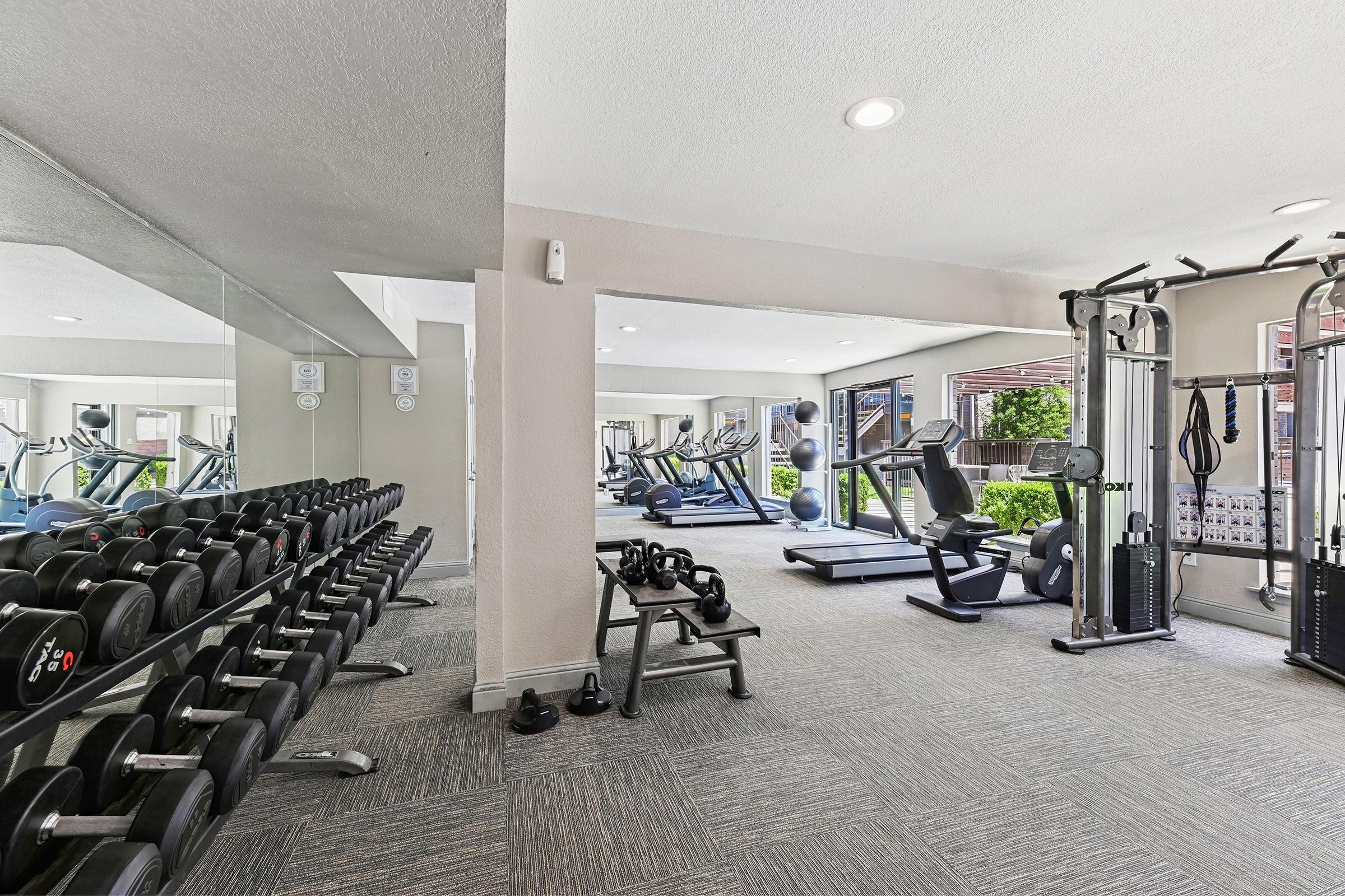 A modern gym interior featuring a variety of exercise equipment. A rack of dumbbells is on the left, while exercise machines, including a weight bench and cable machine, are seen in the background. Large mirrors reflect the equipment, and there are windows providing natural light.