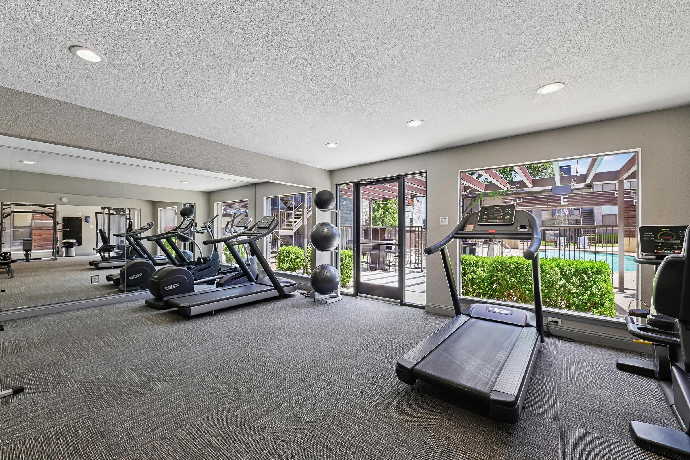 A well-lit gym area featuring cardio equipment like treadmills and ellipticals, surrounded by mirrors. Large windows provide a view of an outdoor space with greenery and a pool. The flooring is carpeted, and the walls are painted in neutral colors for a modern and inviting atmosphere.
