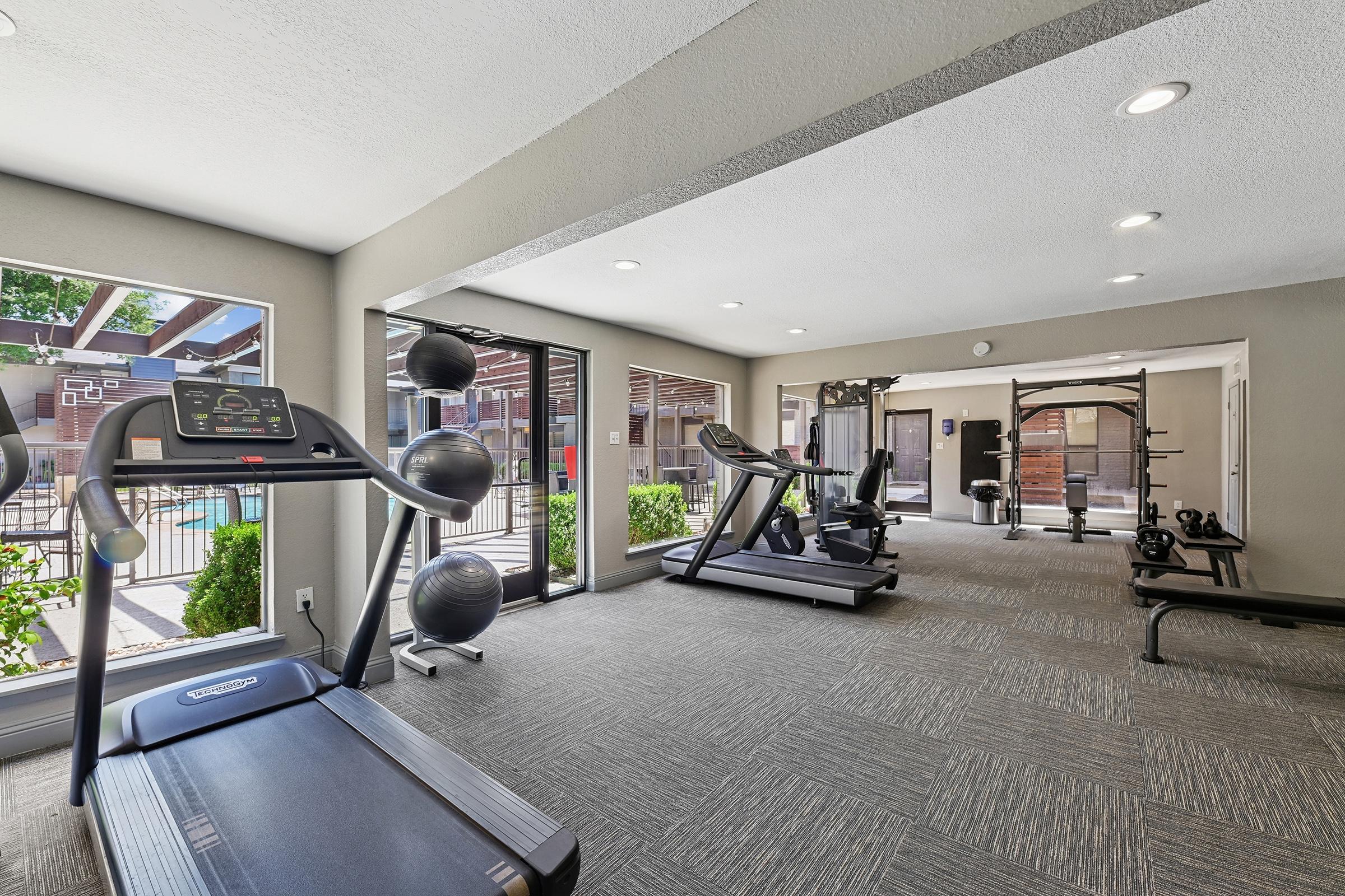 A modern gym interior featuring exercise equipment like a treadmill, elliptical machine, weight benches, and mirrors. Large windows provide natural light and a view of outdoor greenery, creating an inviting workout space. The carpeted floor adds a comfortable ambiance.
