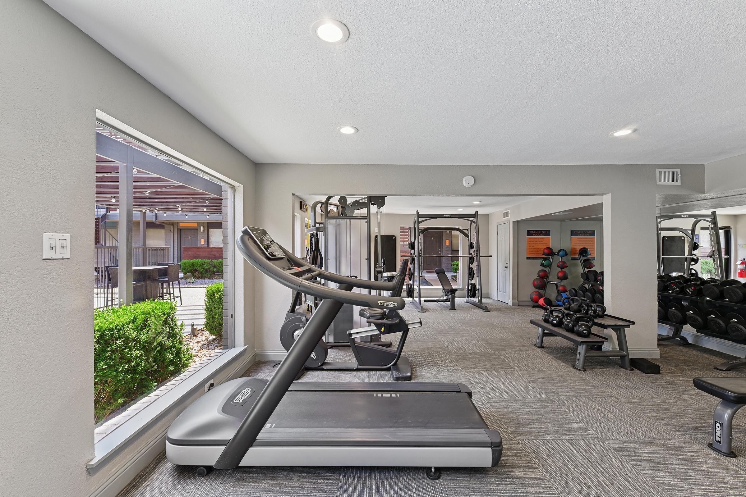 A well-lit fitness room featuring a treadmill in the foreground, surrounded by various workout equipment, including dumbbells and weight machines. Large windows provide natural light and a view of outdoor greenery, enhancing the inviting atmosphere of the gym.