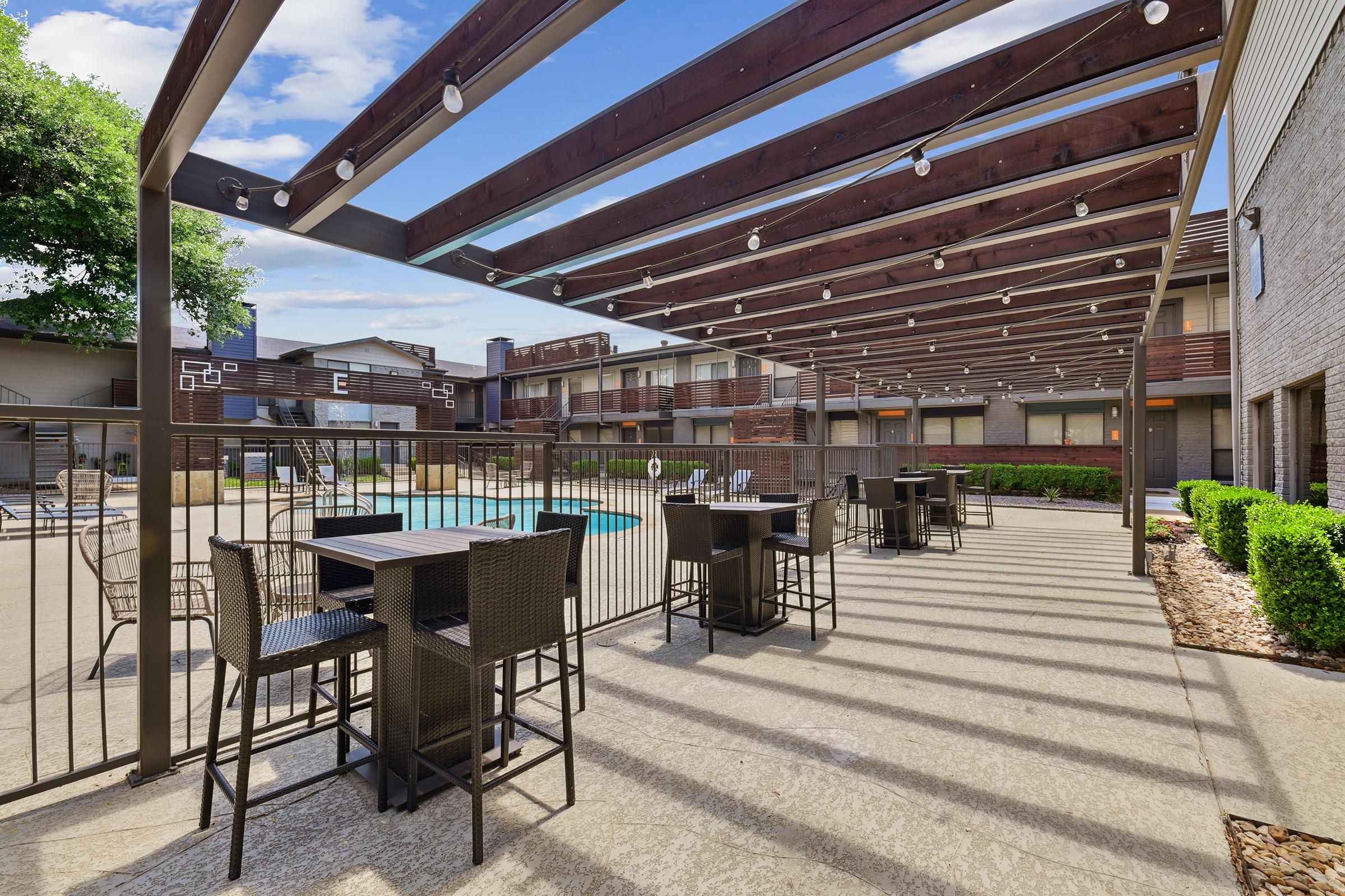 Outdoor area with a wooden pergola featuring string lights, surrounded by a pool and lounge seating. Black patio furniture is arranged in a shaded space, with landscaped greenery and nearby apartment buildings visible, creating a relaxing atmosphere.