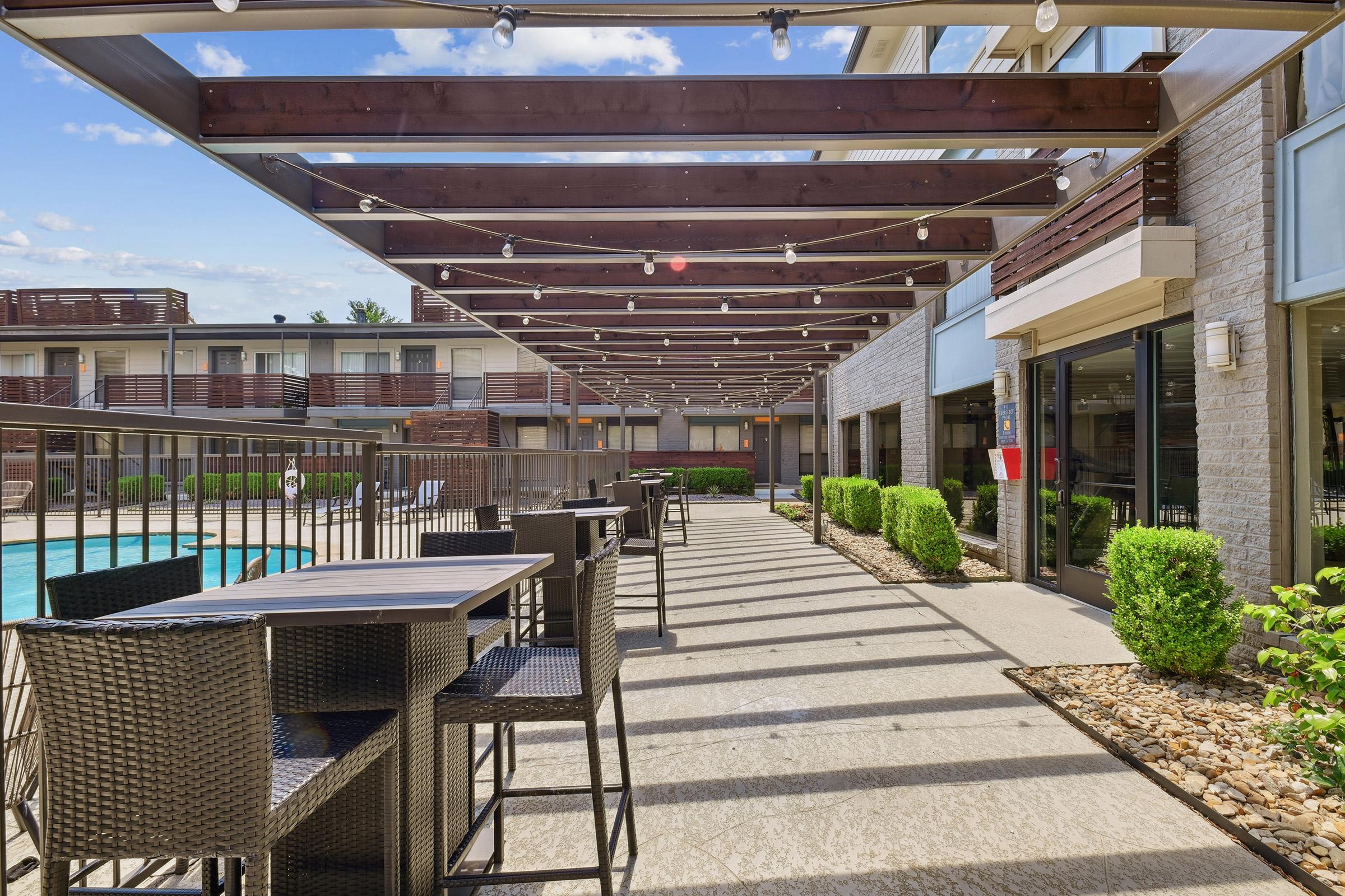 A modern outdoor patio area with shaded seating, featuring dark wicker tables and chairs. The space includes a swimming pool in the background, surrounded by green landscaping, and is under a pergola with string lights. The area has a contemporary, inviting atmosphere.