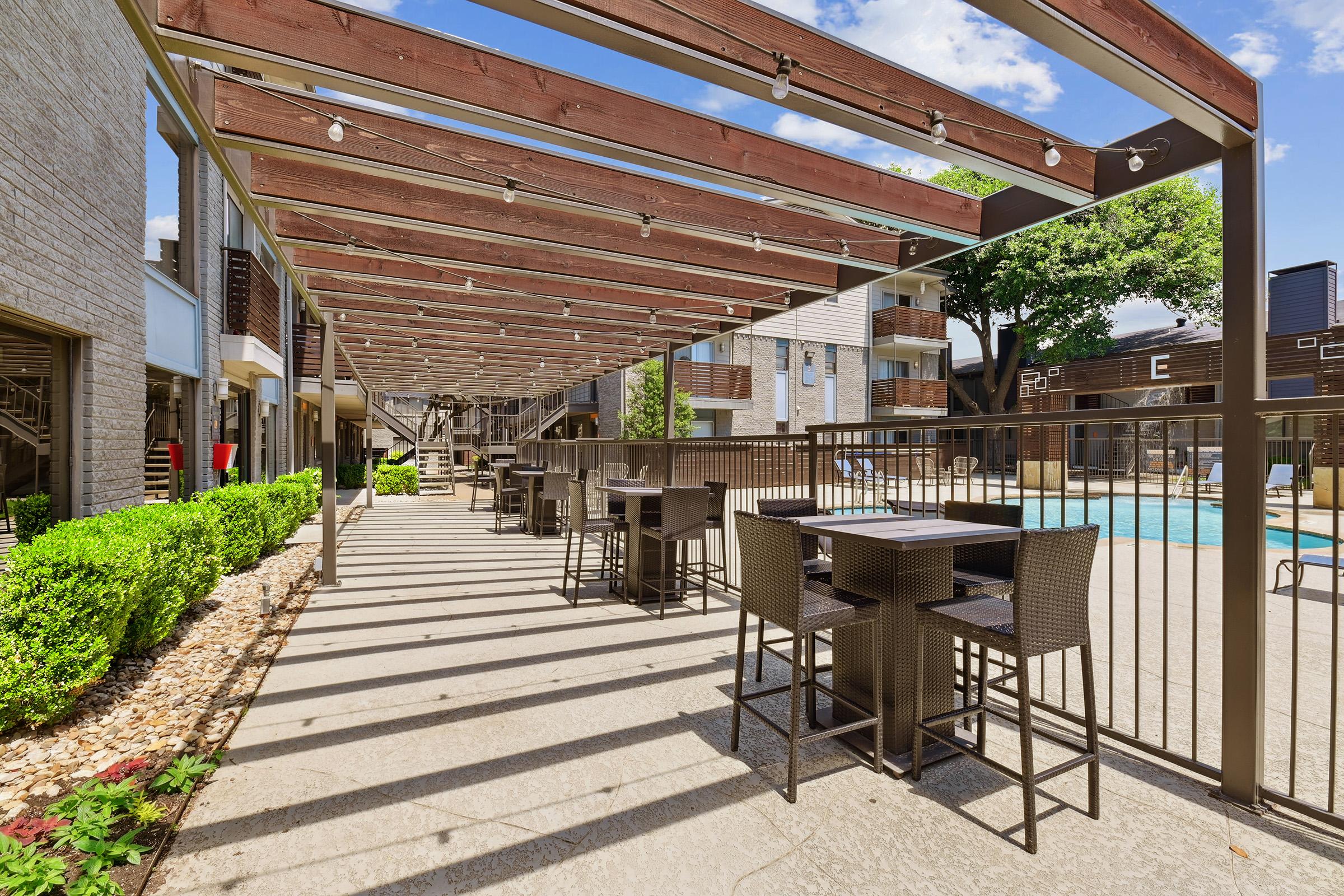 A modern outdoor space featuring a shaded pergola with wooden beams, overlooking a swimming pool. Stylish seating areas with tables and chairs are arranged along the paved pathway, surrounded by well-maintained greenery and plants, creating a relaxing atmosphere.