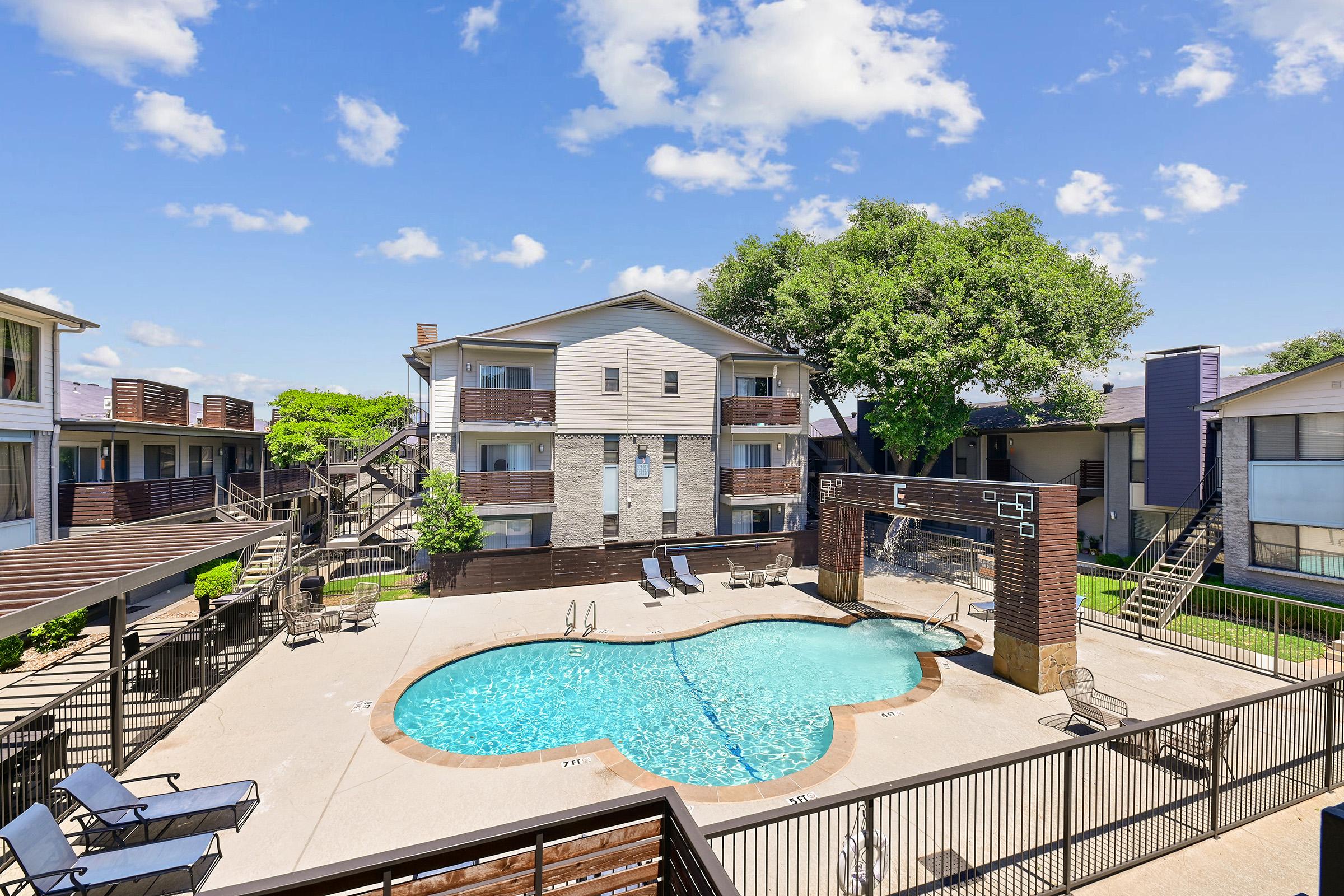 A vibrant apartment complex featuring a refreshing swimming pool at the center, surrounded by lounge chairs. The buildings are two-story with balconies, set against a backdrop of blue skies and fluffy white clouds, providing a welcoming outdoor space for residents.