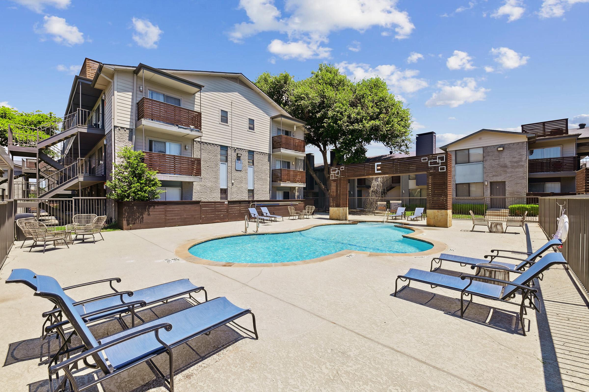 A clear blue swimming pool surrounded by lounge chairs, with two multi-story residential buildings in the background. Lush green trees and a bright blue sky with scattered clouds enhance the inviting atmosphere of this outdoor area.