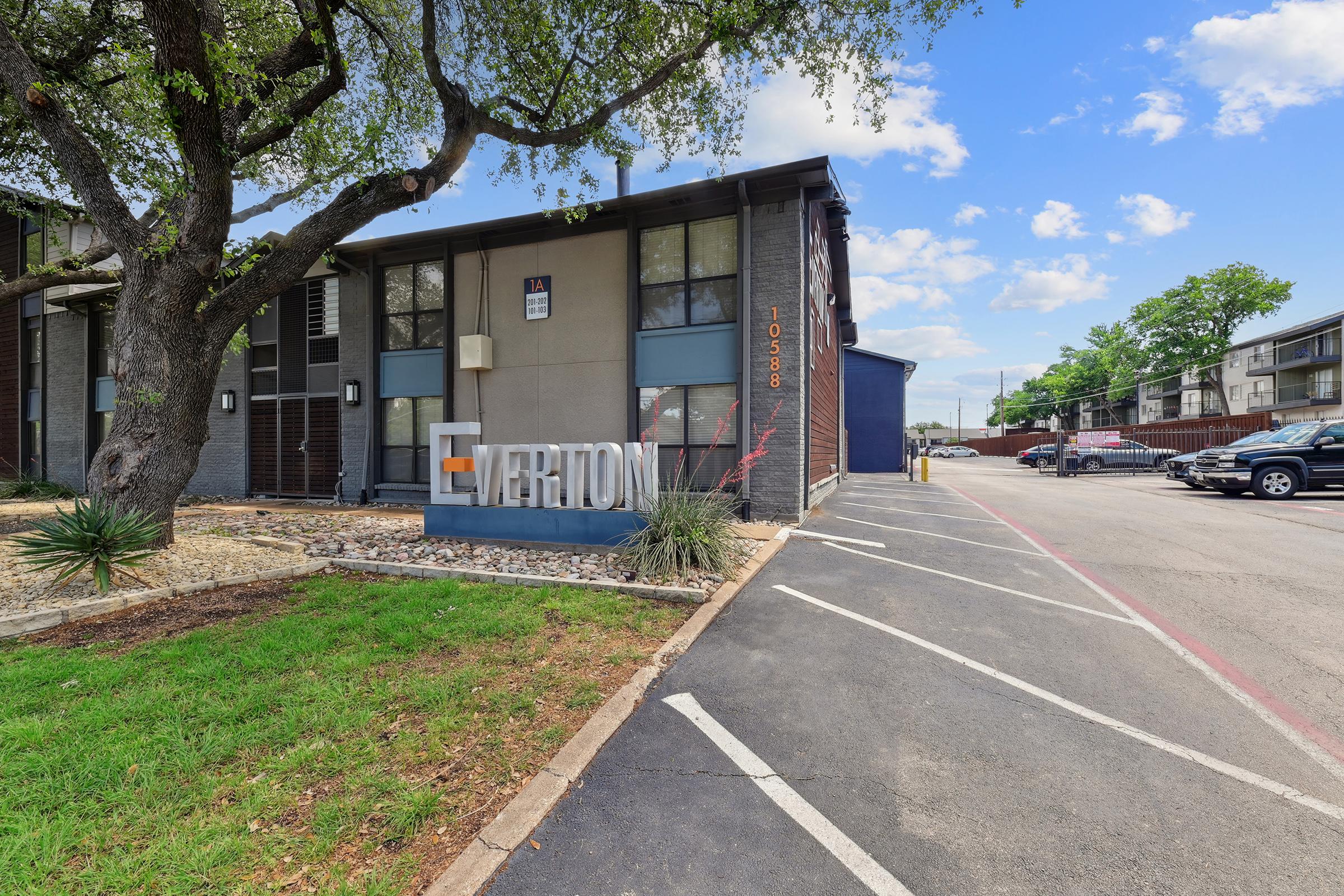 A modern apartment complex entrance featuring a large sign that reads "Everton." The building has a mix of grey and beige exterior with large windows. Surrounding greenery includes trees and shrubs, and there is a paved parking area alongside the building under a clear blue sky with scattered clouds.