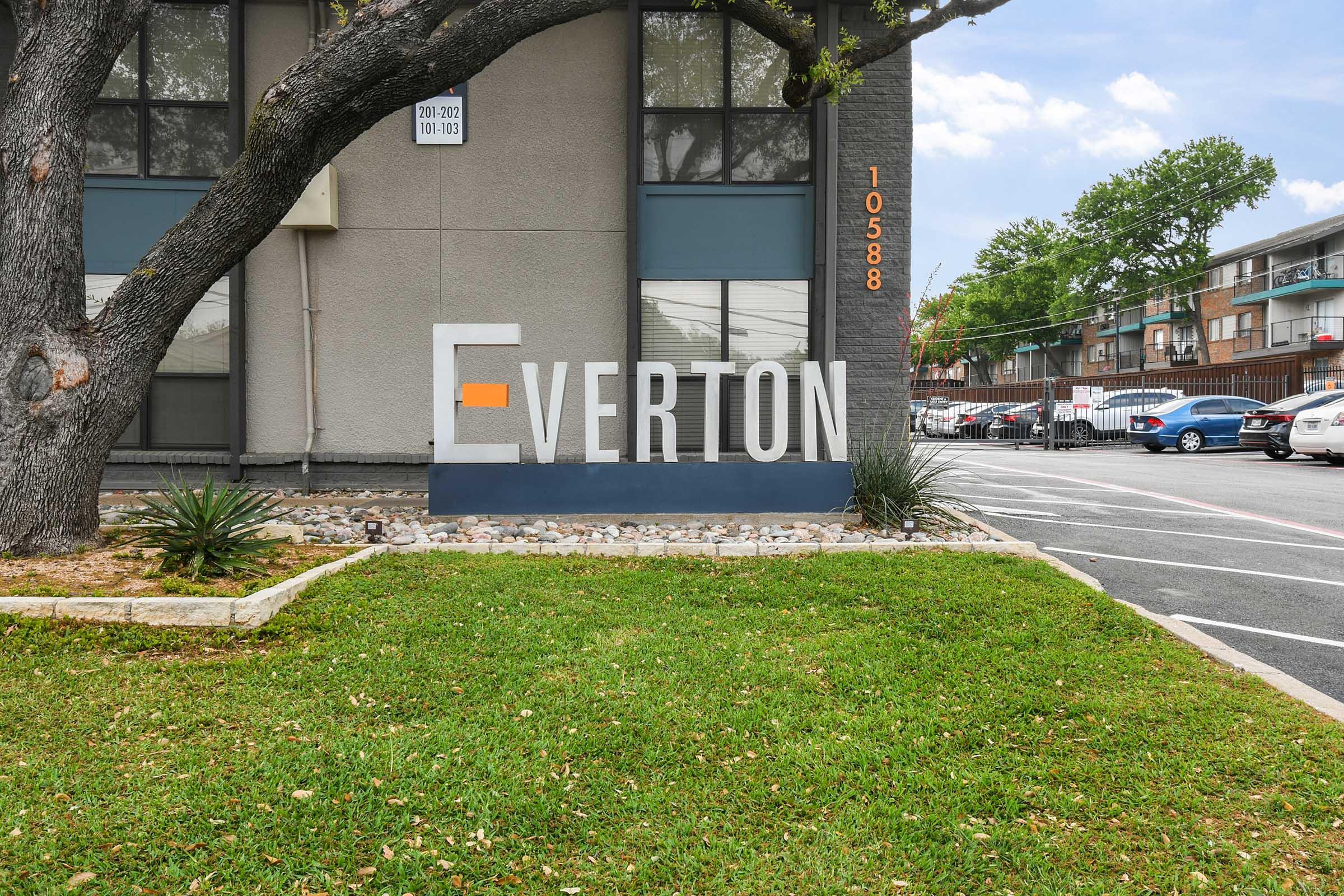 Sign displaying the word "Everton" in bold letters on a building exterior. The sign features an orange accent and is positioned next to a landscaped area with grass and small plants. In the background, there is a parking lot with several cars and additional buildings visible.