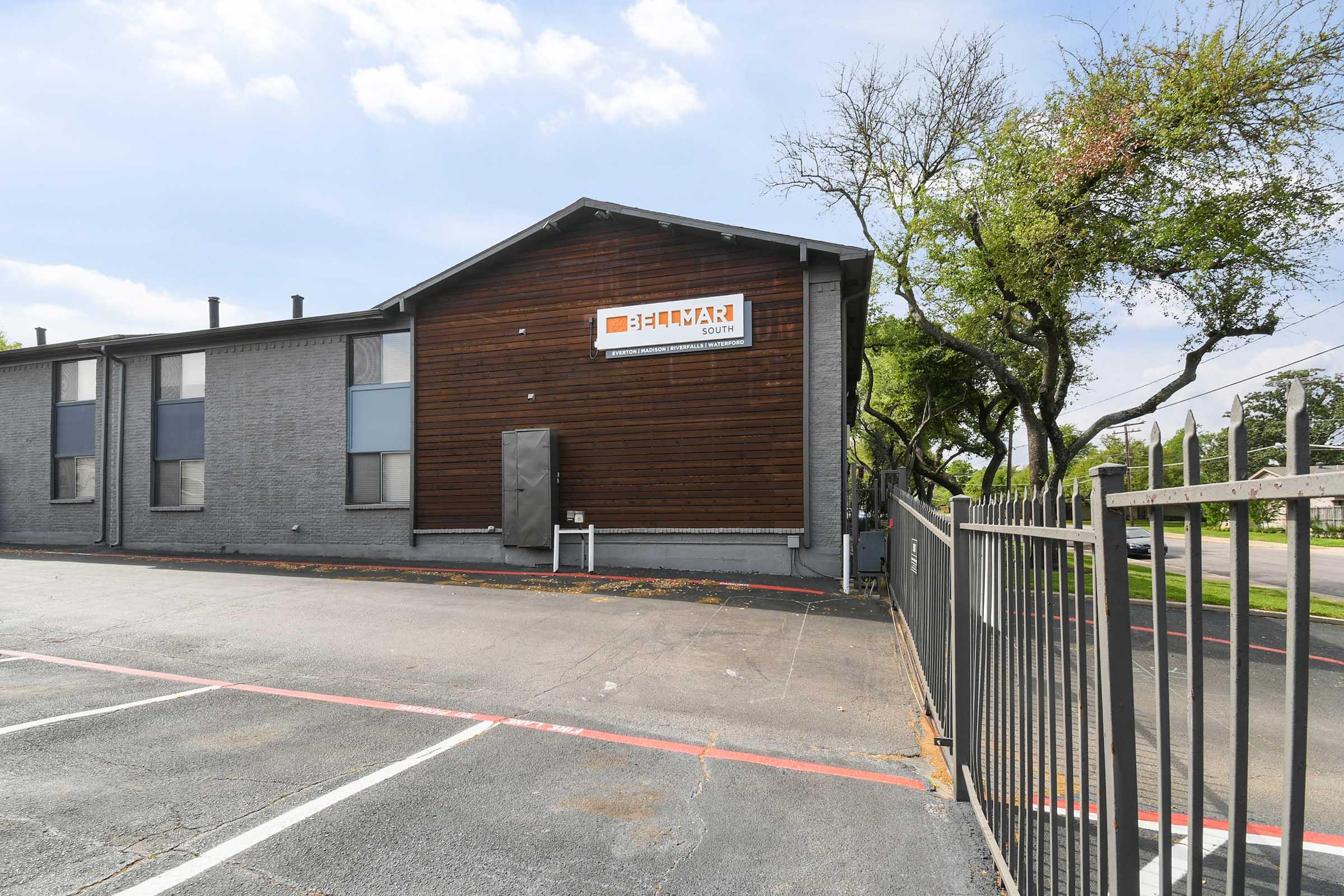 Exterior view of a two-story building featuring a wooden facade and gray walls, with a sign reading "BIMAR" displayed prominently. The image captures the entrance area, a fenced parking lot, and some greenery in the vicinity on a clear day.
