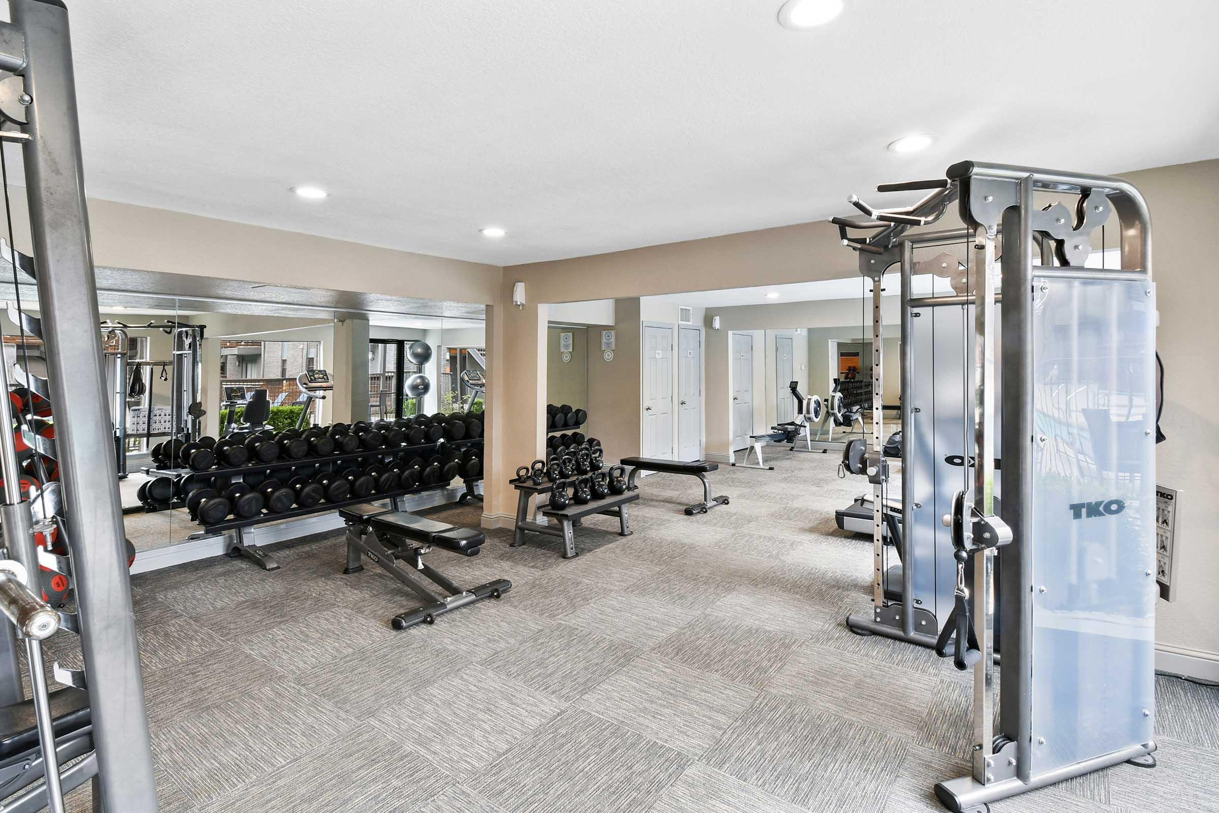 A well-equipped gym interior featuring a variety of weights, including dumbbells on a rack, benches, and multi-station weight machines. The space is bright with overhead lighting and mirrors, providing a clean and modern atmosphere for workouts.