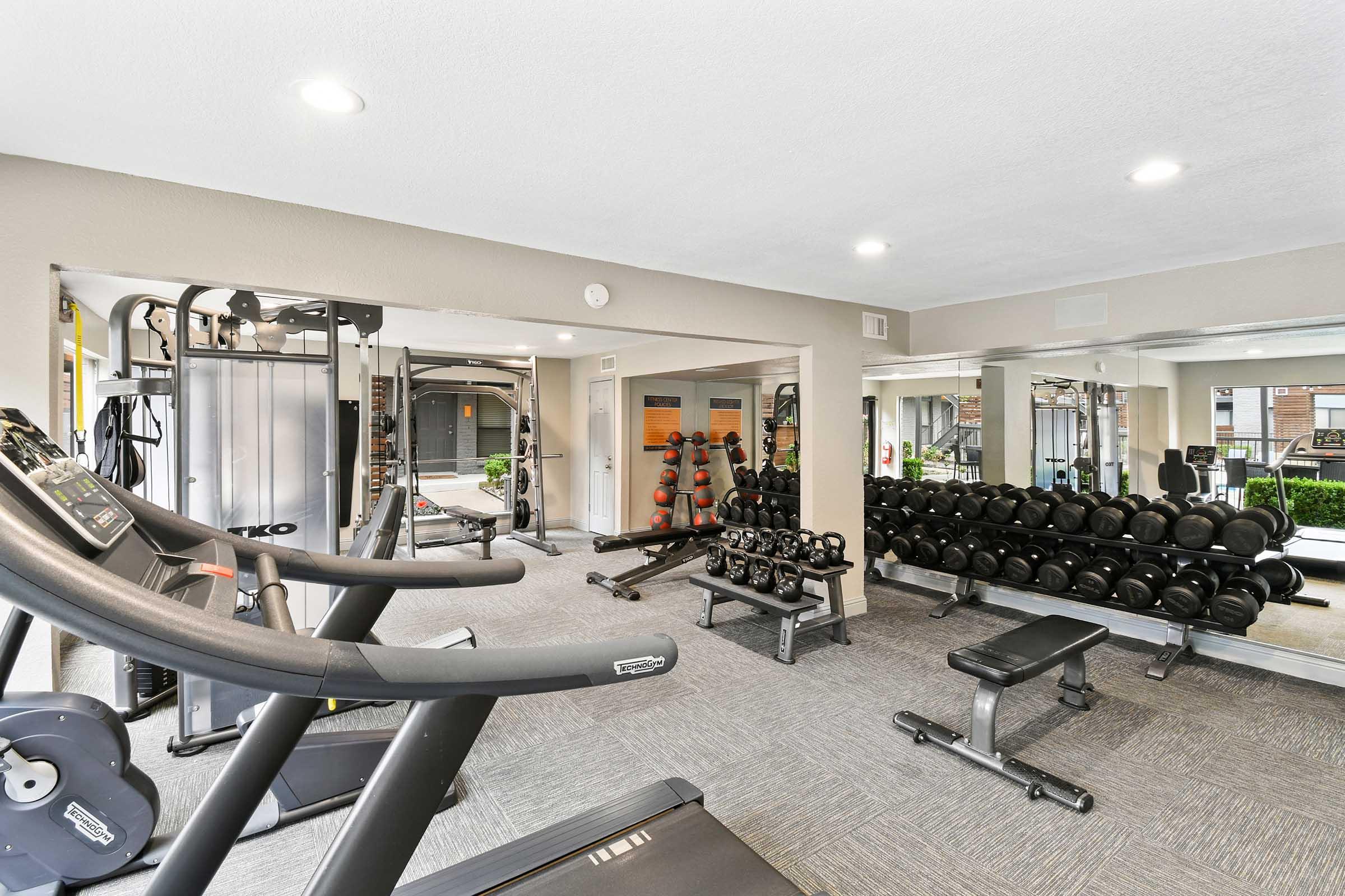 A modern gym interior featuring exercise equipment including treadmills, weight machines, and free weights. The space is well-lit with mirrors on the walls and a clean, organized layout, providing a motivating environment for fitness enthusiasts.