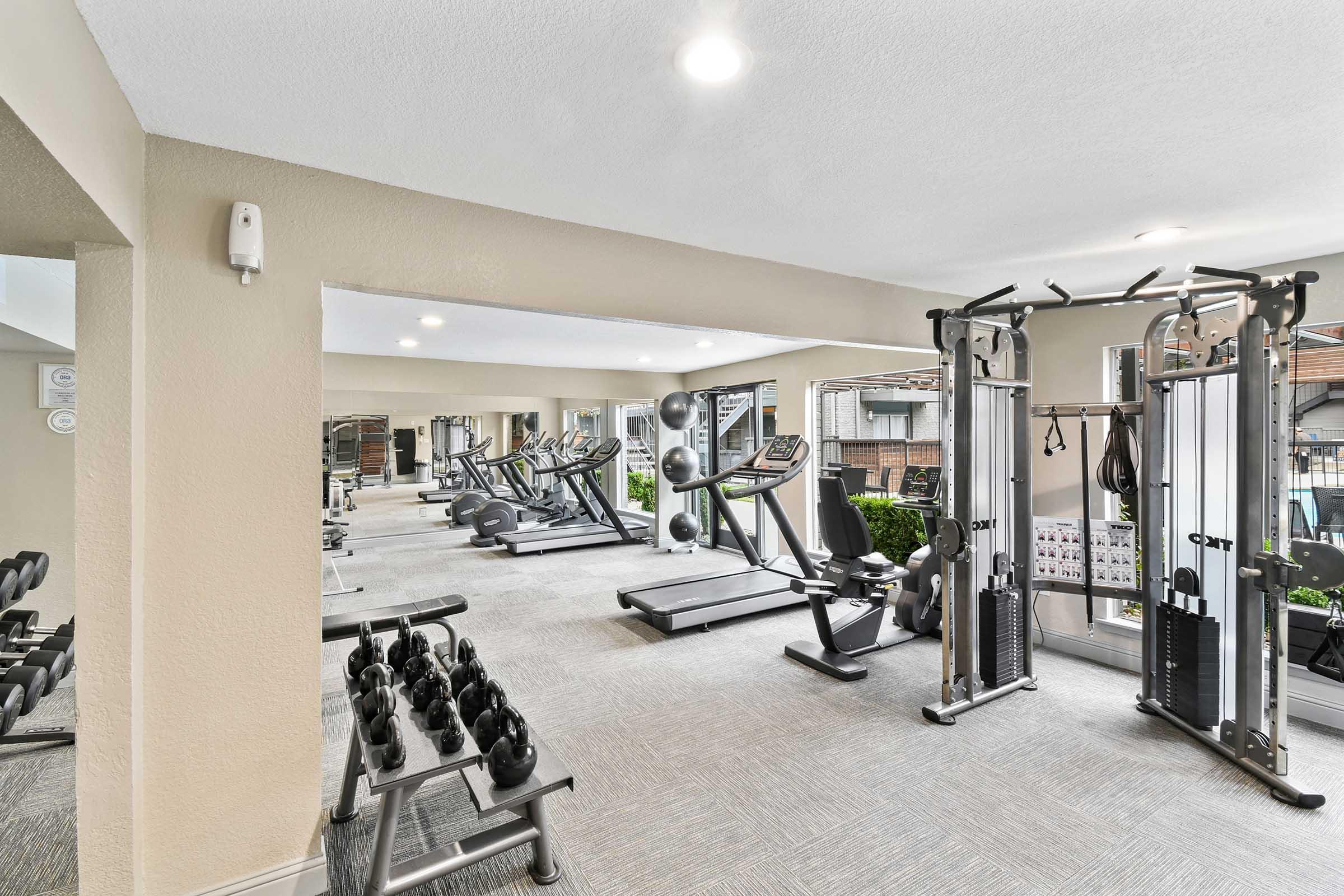 A modern fitness center featuring various workout equipment including cardio machines, weightlifting benches, and free weights. The space is well-lit and designed with a clean, open layout. Large windows provide natural light and a view of greenery.