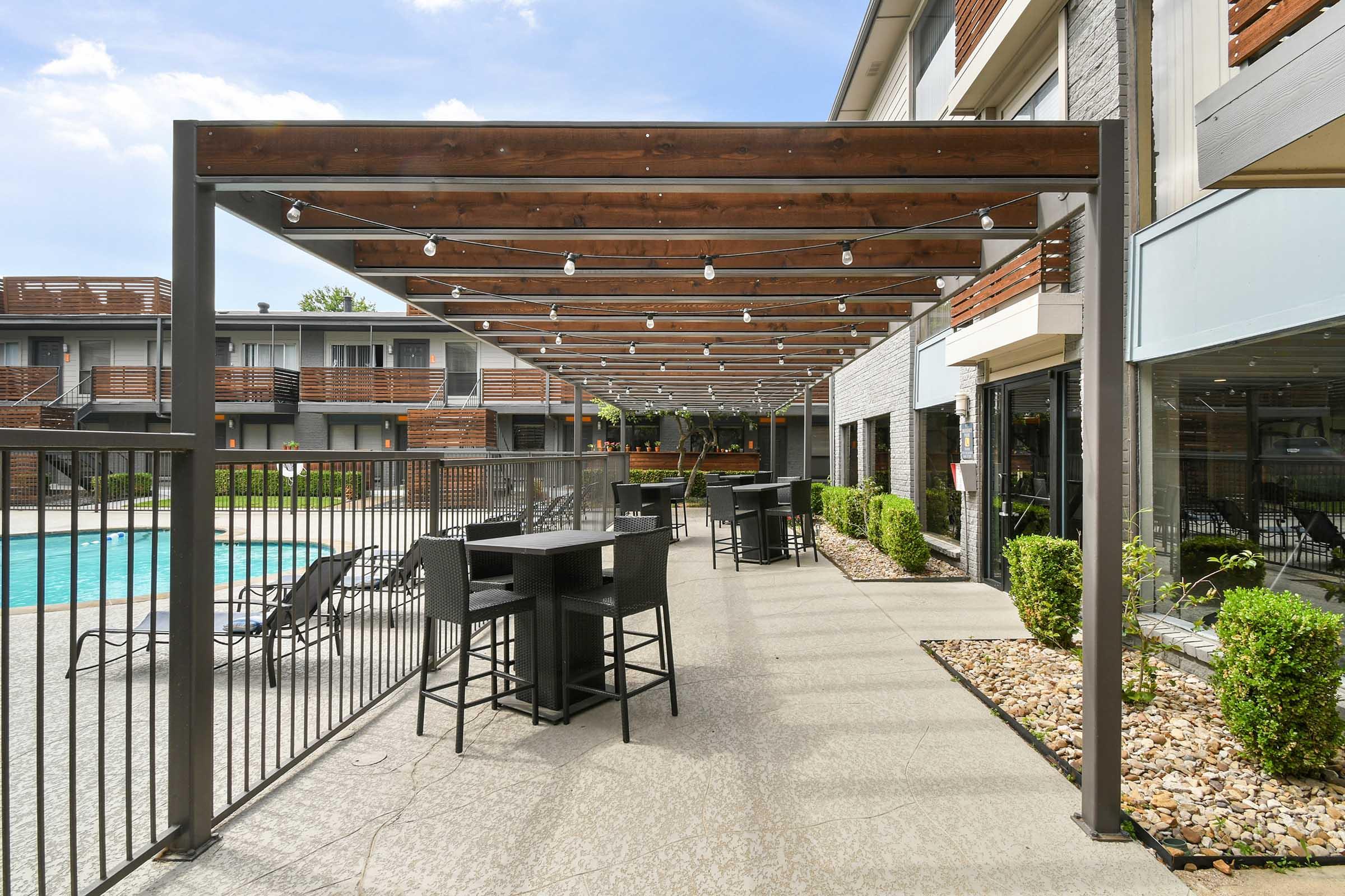 A view of a poolside area featuring a modern pergola with string lights, bar-style seating, and tall black chairs. The outdoor space is flanked by neatly trimmed bushes and leads to a swimming pool in the background, with multi-story buildings visible behind the pool area.