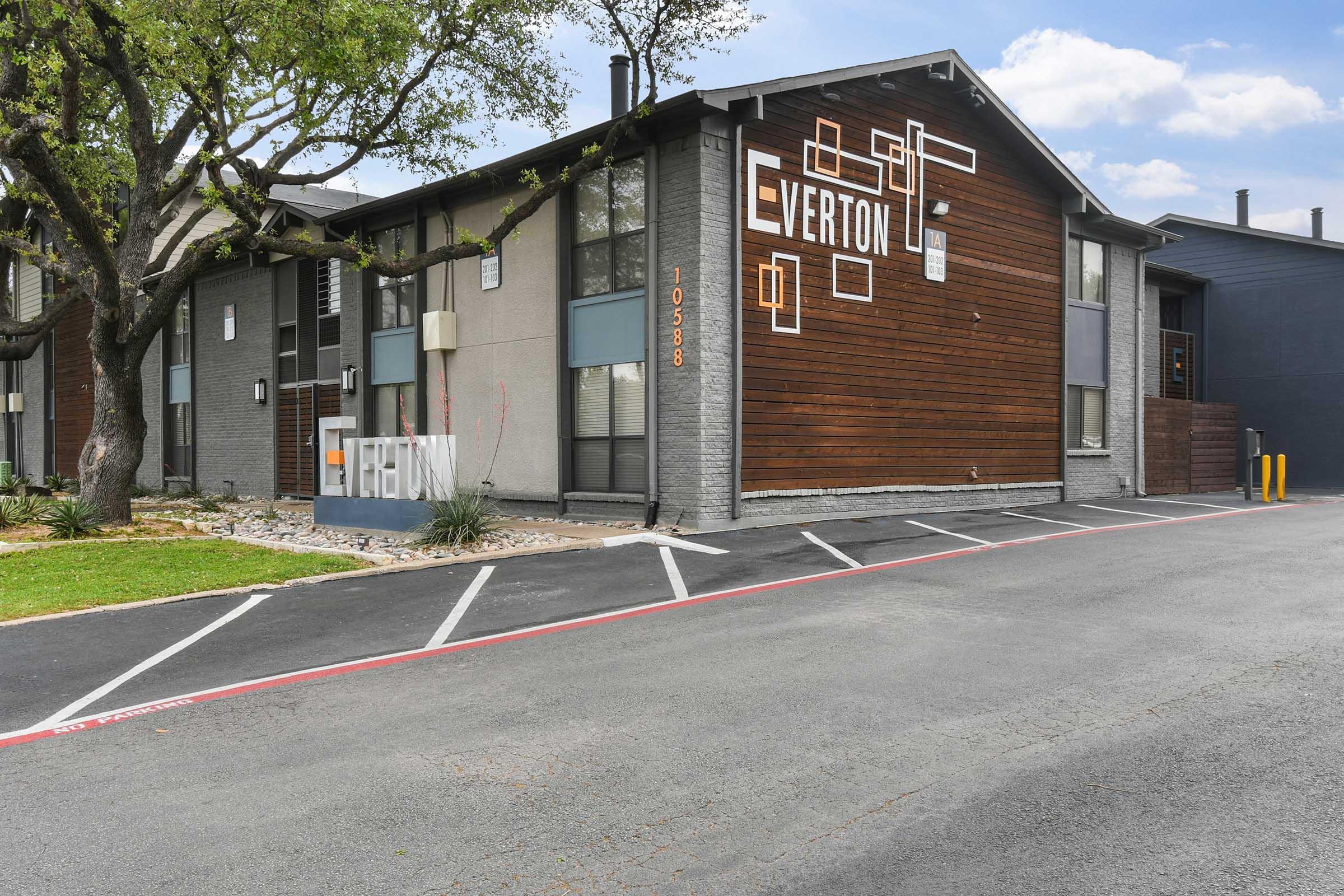 Exterior view of a modern apartment building with a mix of wood and gray siding. The building features a prominent sign that reads "VERTON." In front, a landscaped area with small rocks and greenery, alongside a blacktop parking lot marked with white lines. Clear blue sky in the background.