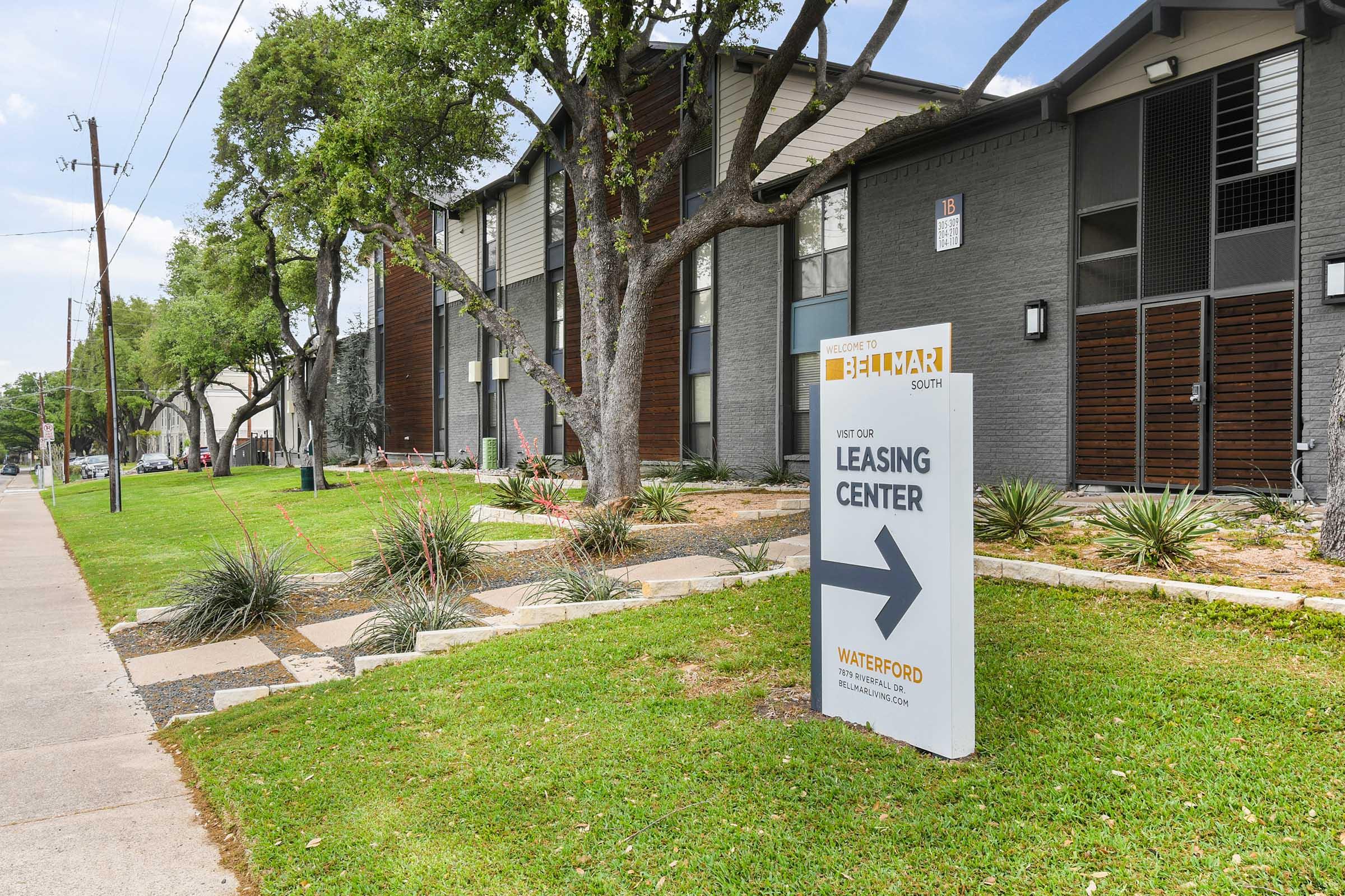 A leasing center sign pointing right, located in front of a modern apartment building surrounded by grass and landscaping. Trees line the walkway, and the building features a mix of light and dark exterior materials.
