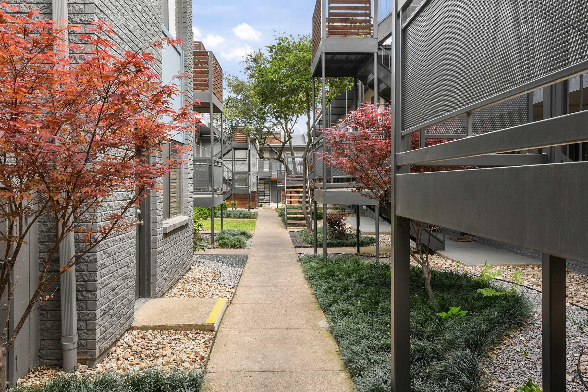 A serene walkway between modern apartment buildings, featuring neatly landscaped green lawns and ornamental plants. Red-leafed bushes line the path, while metal staircases lead to upper levels. The sky is partly cloudy, creating a peaceful atmosphere.