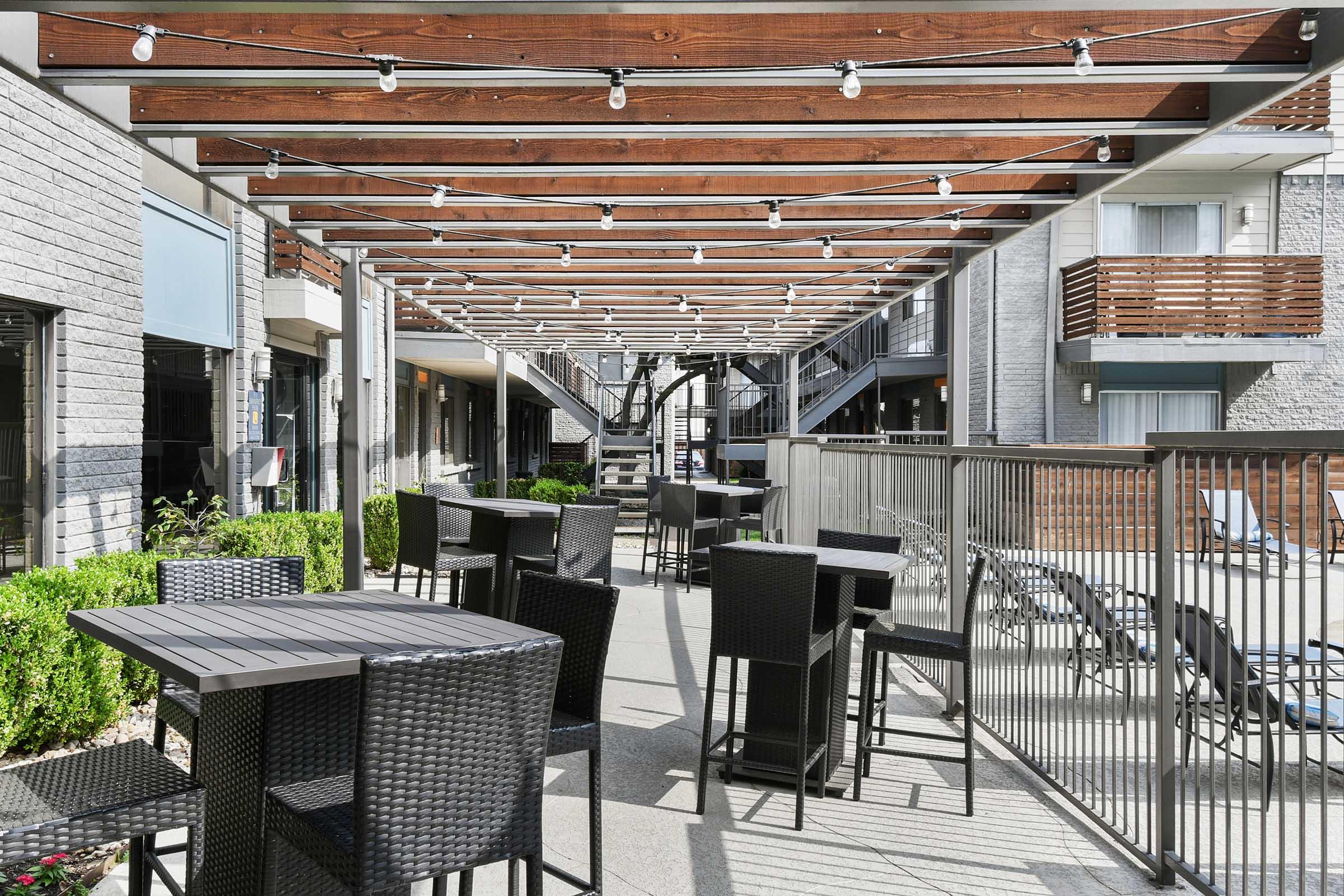 Outdoor dining area featuring neatly arranged black tables and chairs beneath a wooden pergola with string lights. The space is surrounded by greenery and modern buildings, creating a cozy atmosphere for guests. A spiral staircase is visible in the background, adding architectural interest.