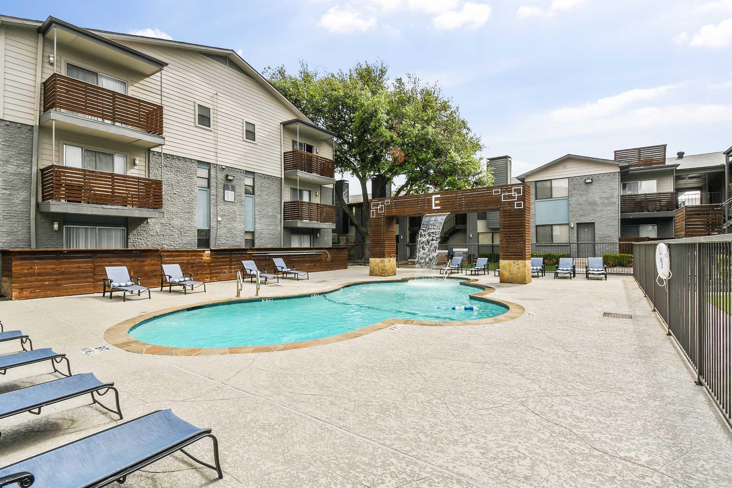 A clear swimming pool surrounded by lounge chairs, with two apartment buildings in the background. The pool features a stone waterfall and is enclosed by a fence. Trees provide shade next to the area, under a bright blue sky with a few clouds.
