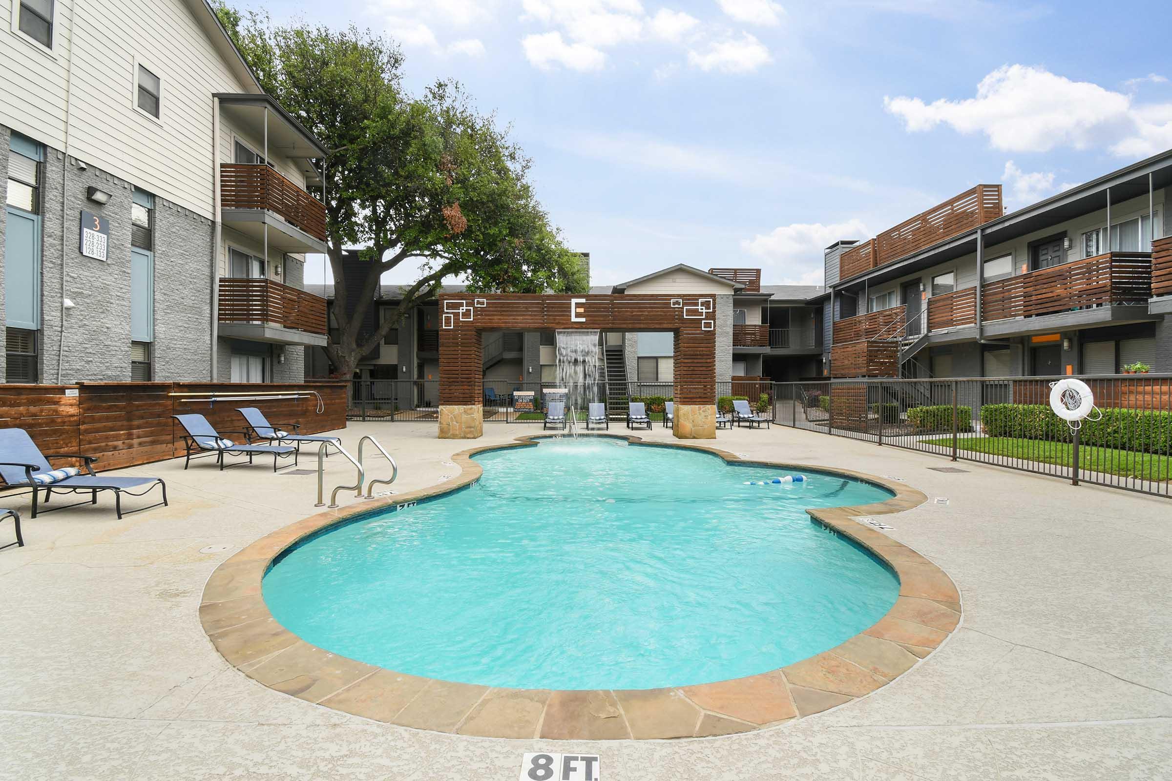 A swimming pool surrounded by lounge chairs, with an apartment complex in the background. The pool has a unique shape and features a decorative entryway marked with the letter "E." Lush green trees are visible, and the sky is partly cloudy.