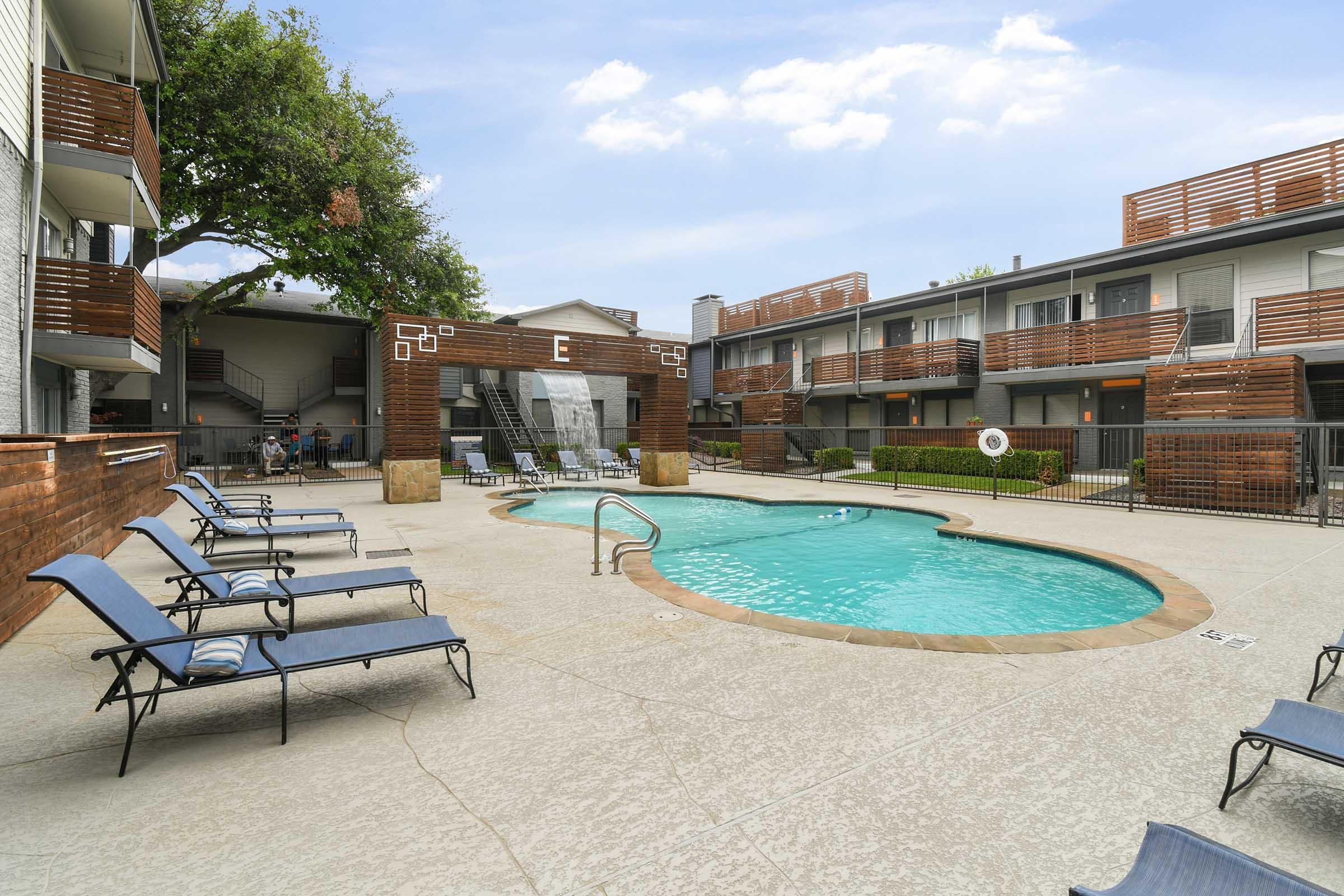 A swimming pool surrounded by lounge chairs in a courtyard setting. The pool area features a modern design with wood accents and landscaping. Apartment buildings are visible in the background, creating a relaxed atmosphere. A clear blue sky is overhead.