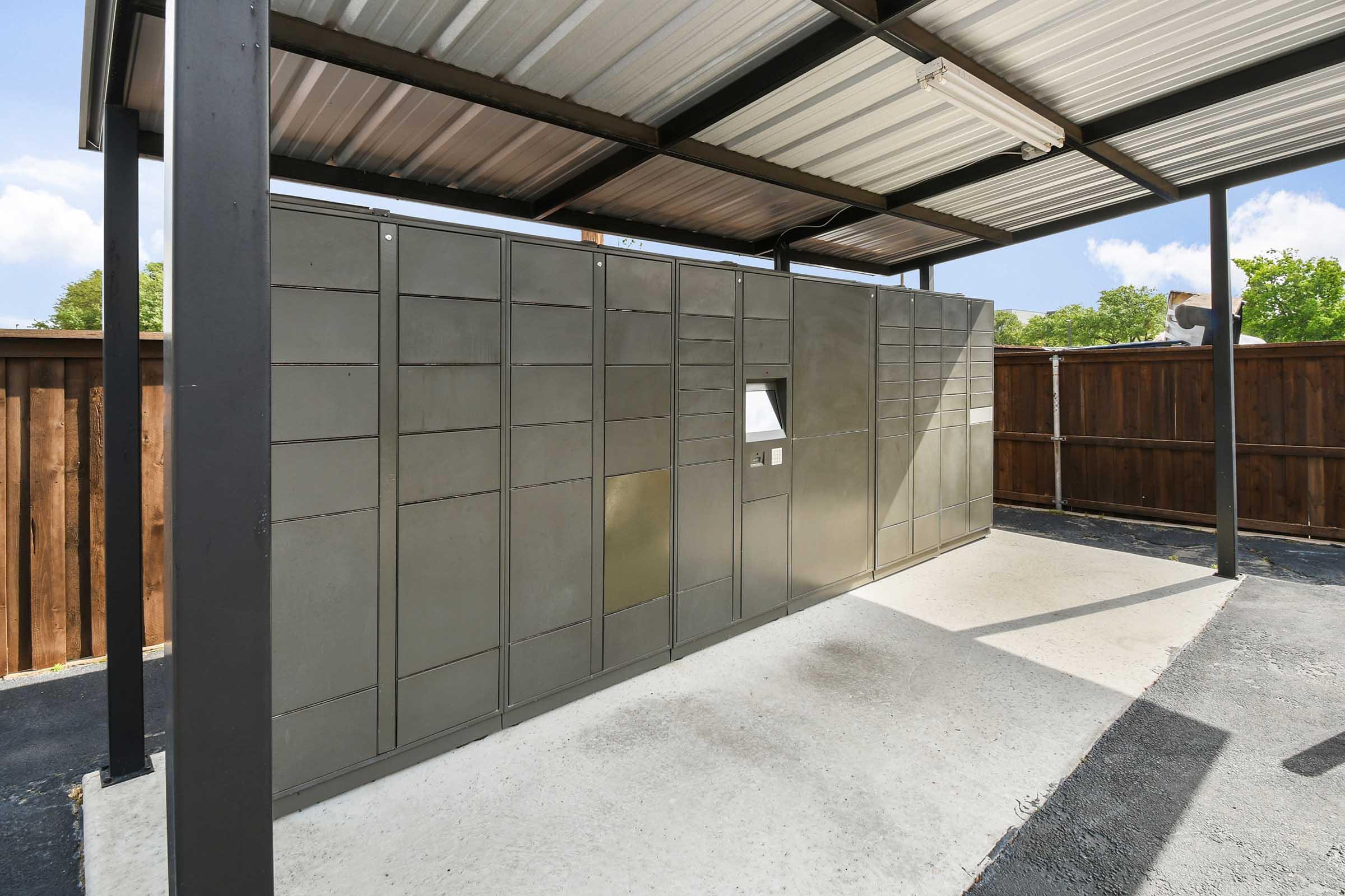 A row of metal mailboxes housed under a covered shelter, situated in a concrete area. The backdrop features a wooden fence and greenery, with blue skies visible above. The design is utilitarian, providing storage for mail and packages in a secure outdoor space.