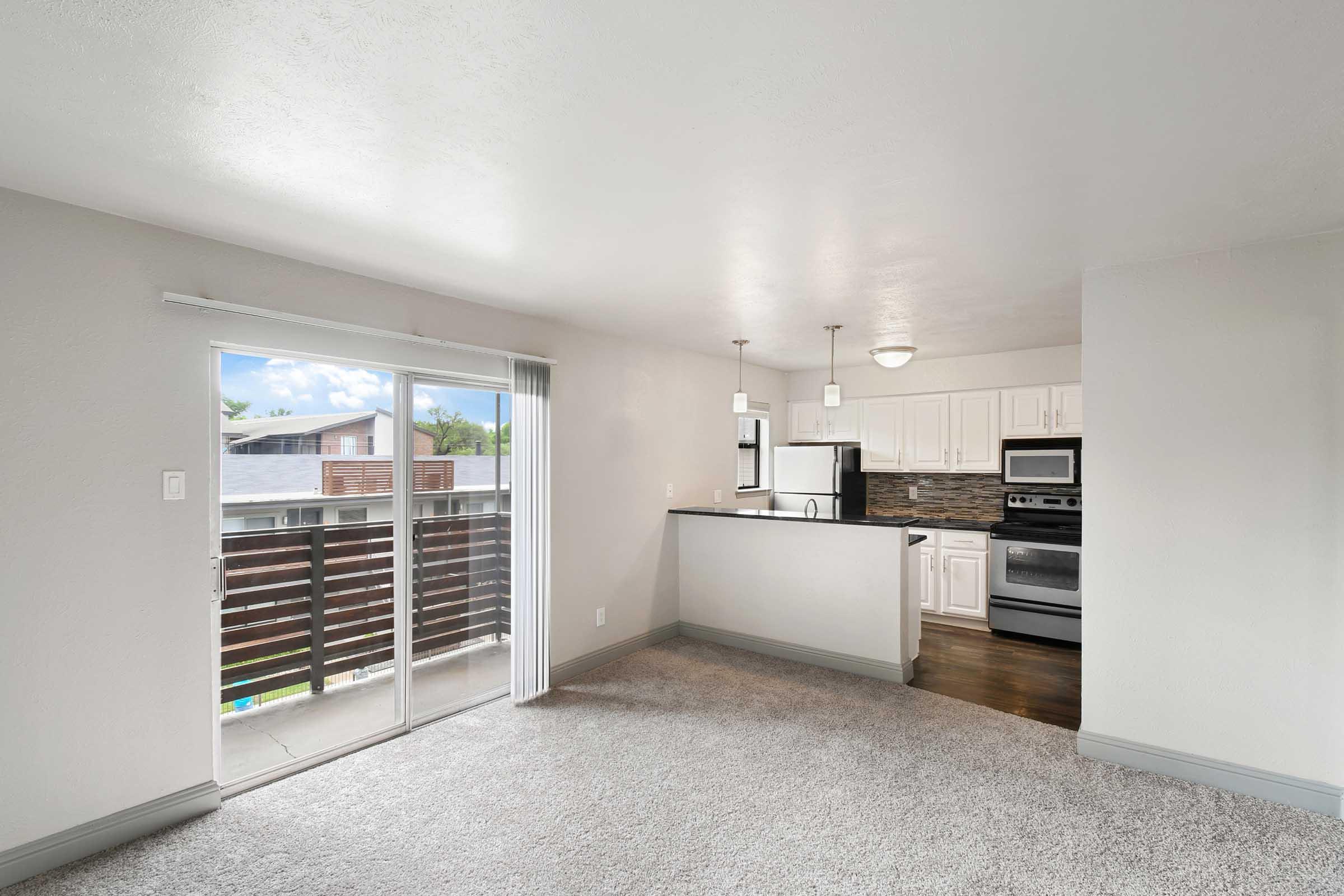 A modern, bright living area featuring plush carpet flooring and an adjoining kitchen. The kitchen has white cabinets, stainless steel appliances, and a breakfast bar. A sliding glass door leads to a small balcony with outdoor views. Natural light fills the space through windows.