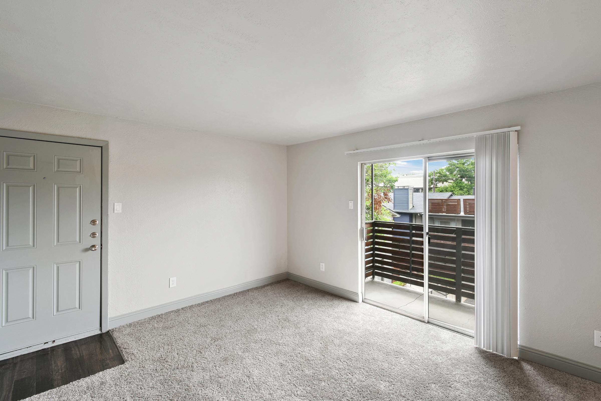 A light-filled room with beige walls and a carpeted floor. There's a gray front door on the left and a large sliding glass door on the right leading to a small balcony. Natural light streams in, highlighting the space's potential for furnishings and decor.