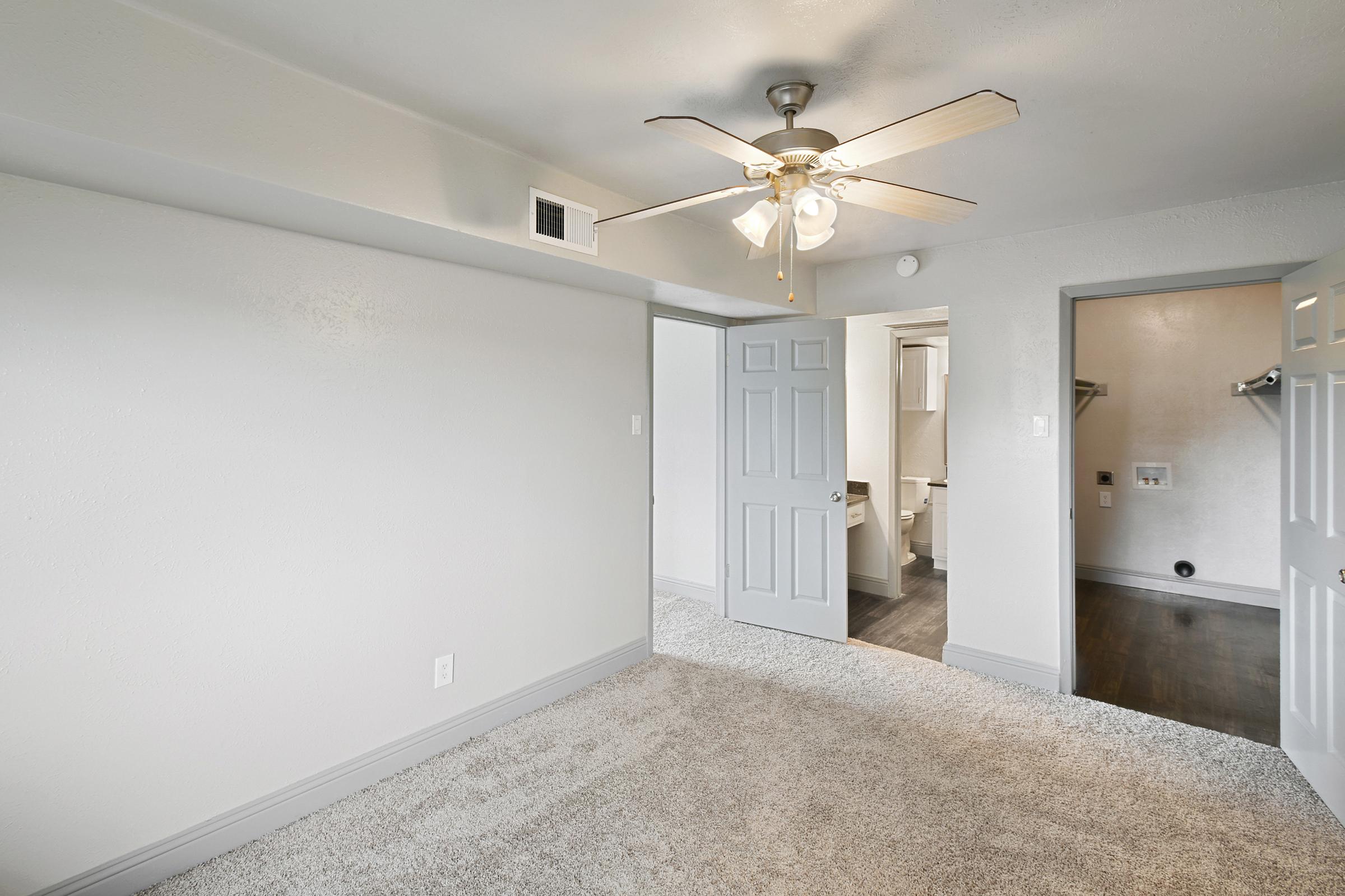 A view of a spacious, well-lit room with light gray walls and carpeted flooring. It features a ceiling fan with five blades, an open door leading to an adjacent room with a closet, and a doorway to a bathroom. The overall aesthetic is modern and inviting.