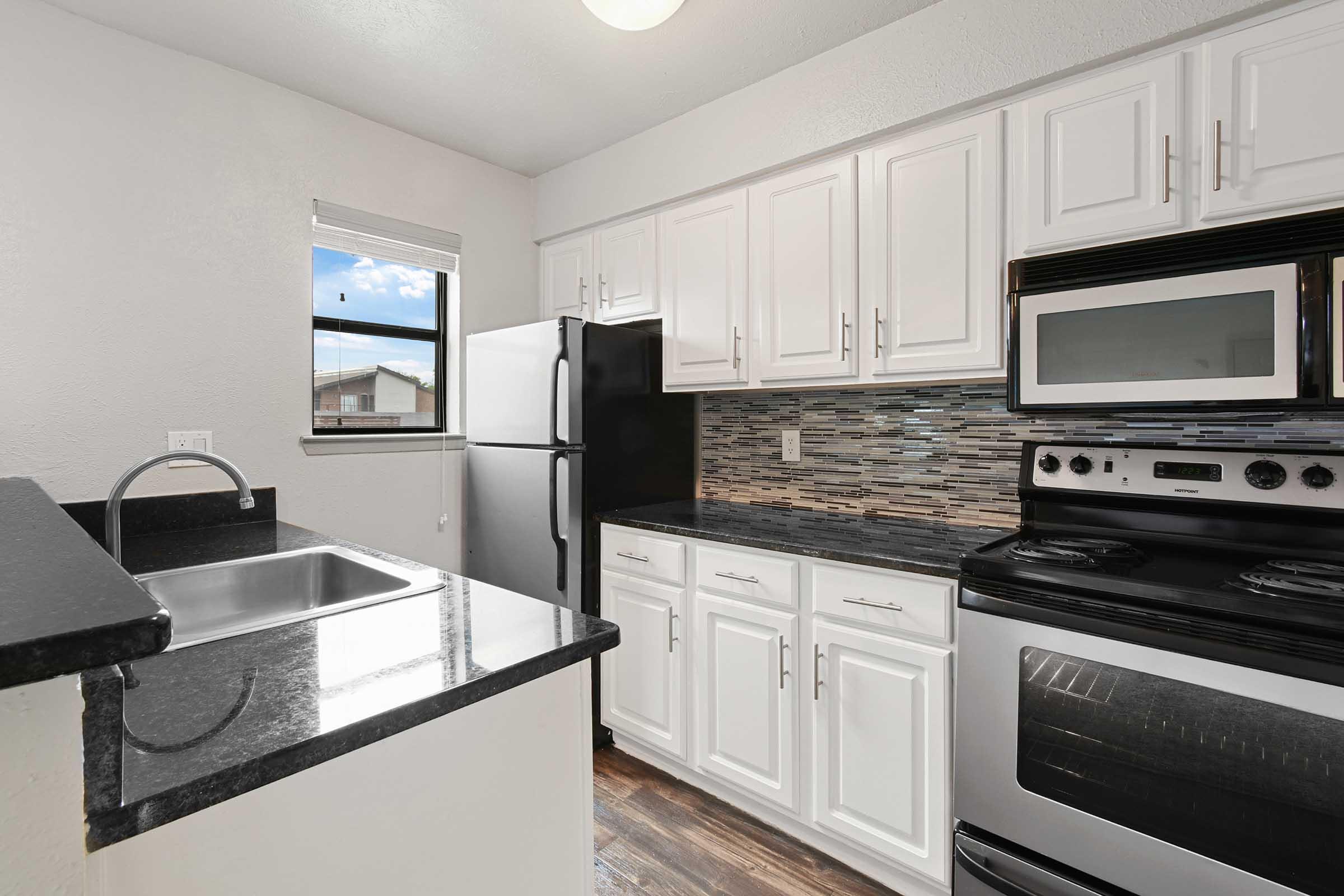 A modern kitchen featuring white cabinets, dark granite countertops, a stainless steel sink, and a window letting in natural light. Appliances include a black refrigerator, a built-in microwave, and an electric stove and oven. The backsplash is made of decorative tile, enhancing the overall aesthetic.