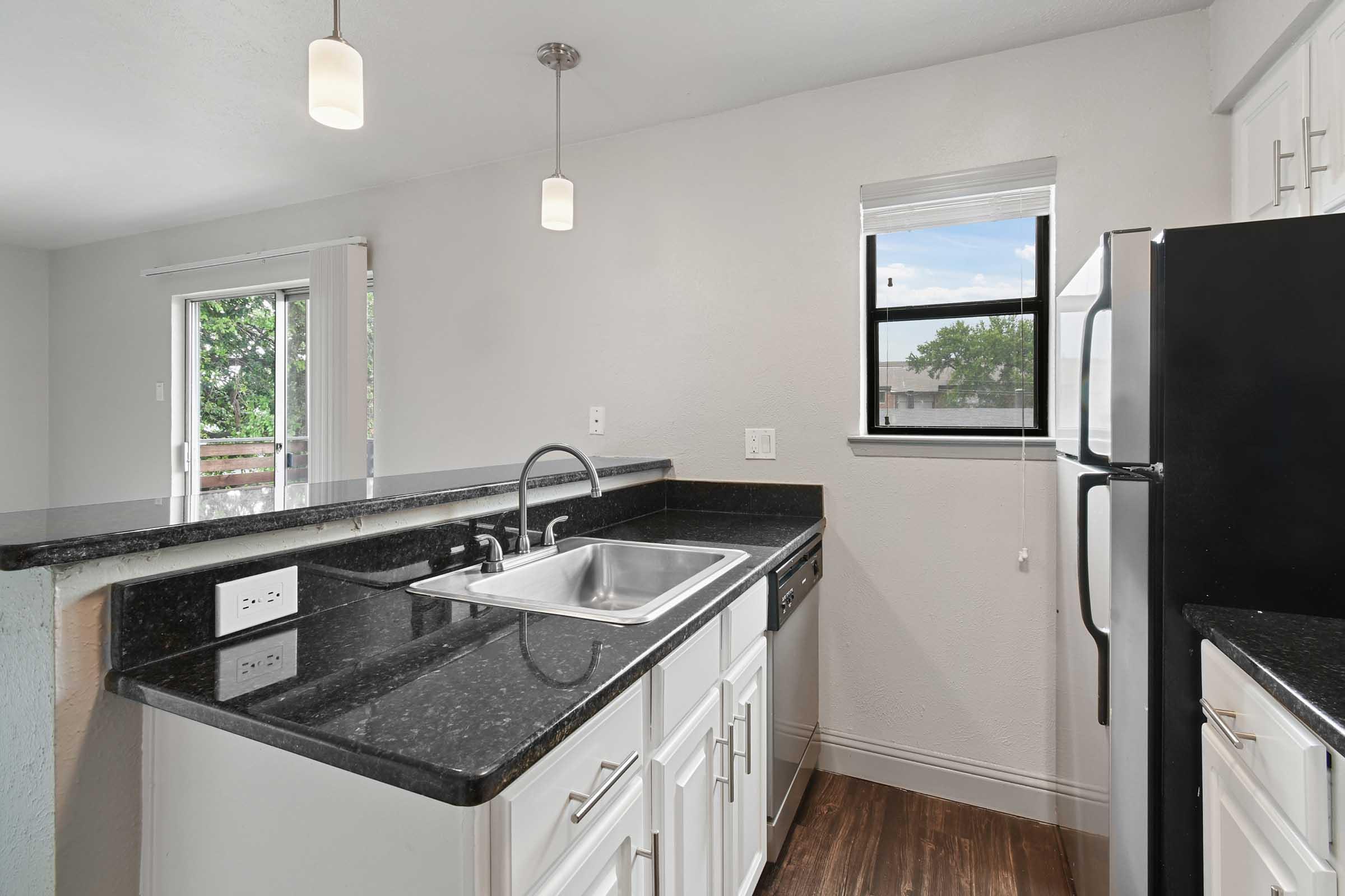 Modern kitchen featuring white cabinetry, dark granite countertops, and stainless steel appliances. A sink is situated beneath a window overlooking greenery. Two pendant lights hang from the ceiling, and a doorway leads to a balcony. The flooring is a warm brown tone, enhancing the contemporary look.