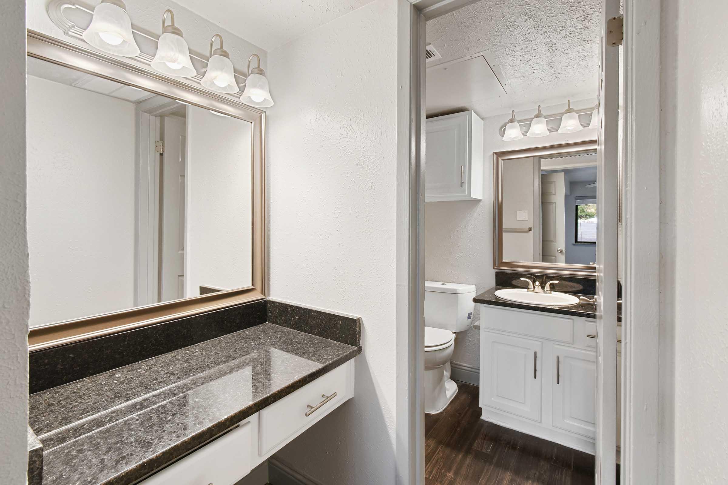 A modern bathroom featuring two mirrors with light fixtures above, a double sink on a dark granite countertop, a toilet in the corner, and a doorway leading to another room. The walls are painted white, and the floor has a dark wood finish. 