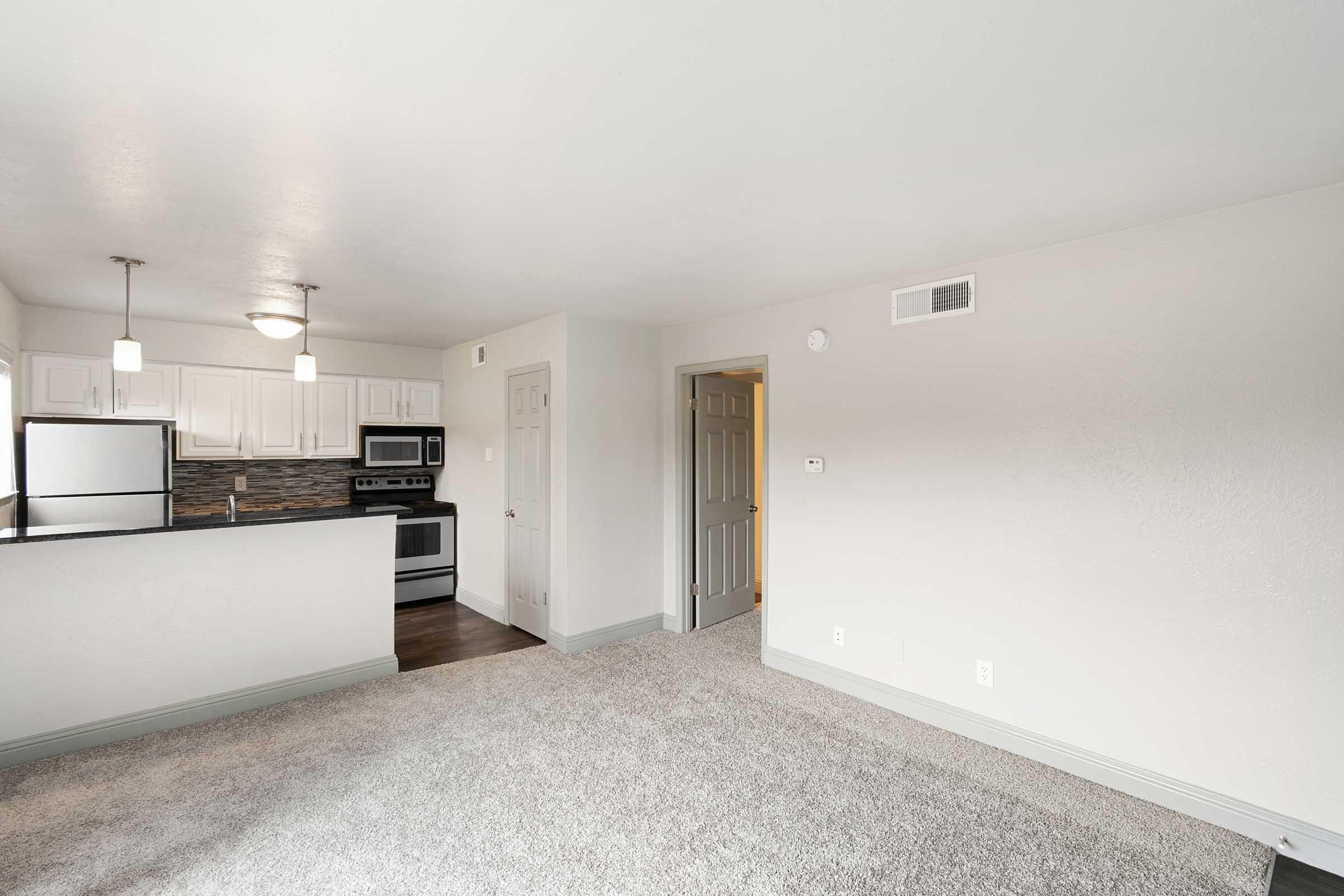 A bright, modern kitchen and living area featuring gray carpeting, white cabinetry, stainless steel appliances, and a textured backsplash. A doorway leads to another room, while the space is well-lit with recessed lighting and a pendant light hanging over the kitchen counter.