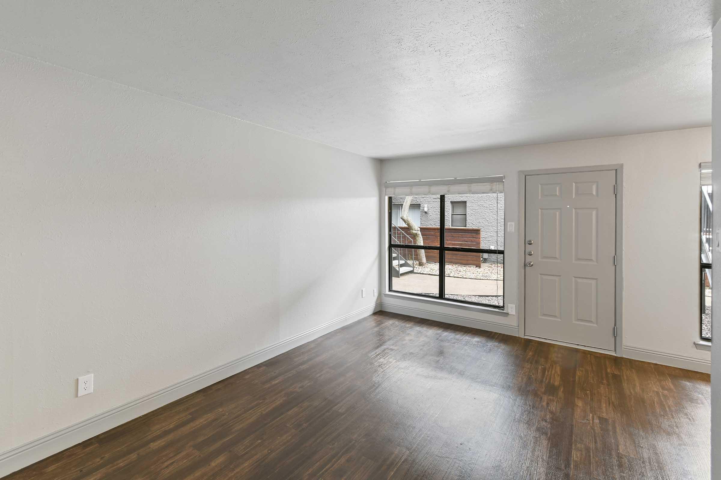 Empty room with light-colored walls and dark wooden flooring. A large window on the right side lets in natural light, revealing a view outside. The door is positioned on the left, and the room appears spacious and bright.