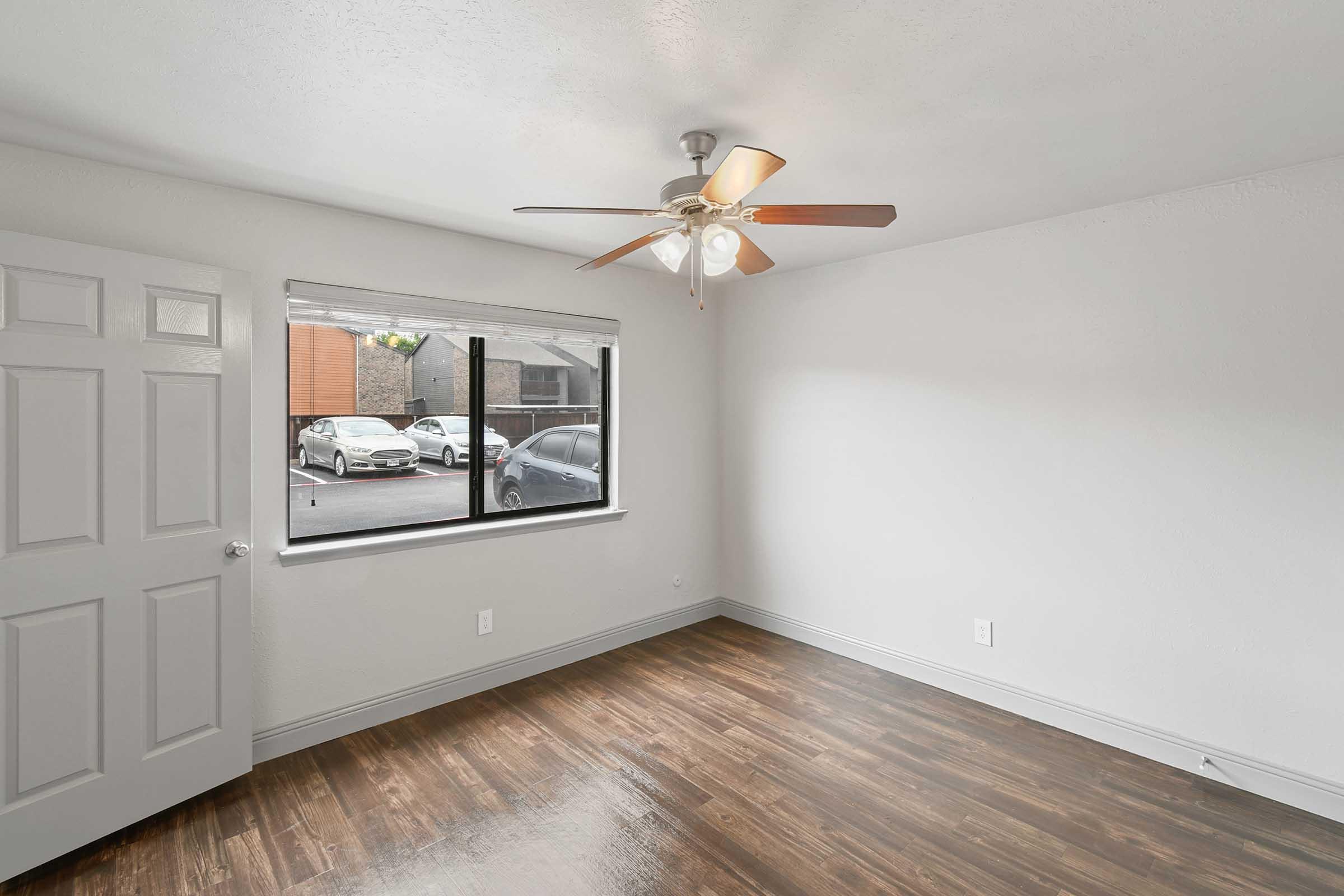 A bright, empty room featuring a ceiling fan and a large window with a view of parked cars outside. The floor is made of wood-like laminate, and the walls are painted white. A closed door is visible on the left side of the image.