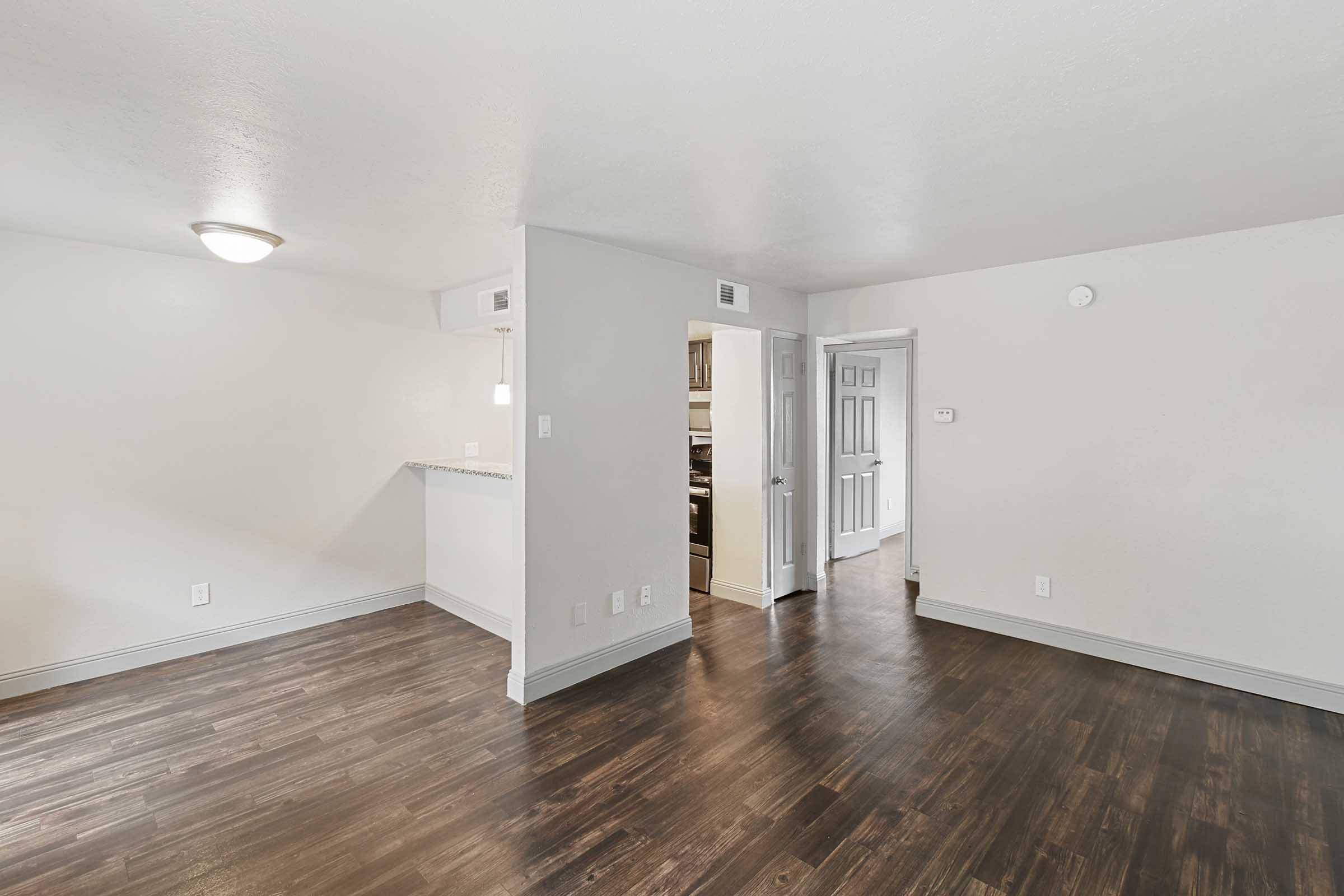 Bright and spacious interior of a modern apartment featuring light walls, dark hardwood flooring, and a door leading to a kitchen area. The layout includes a small entry space with doorways to different rooms, creating an open and inviting atmosphere. Natural light enhances the clean design.