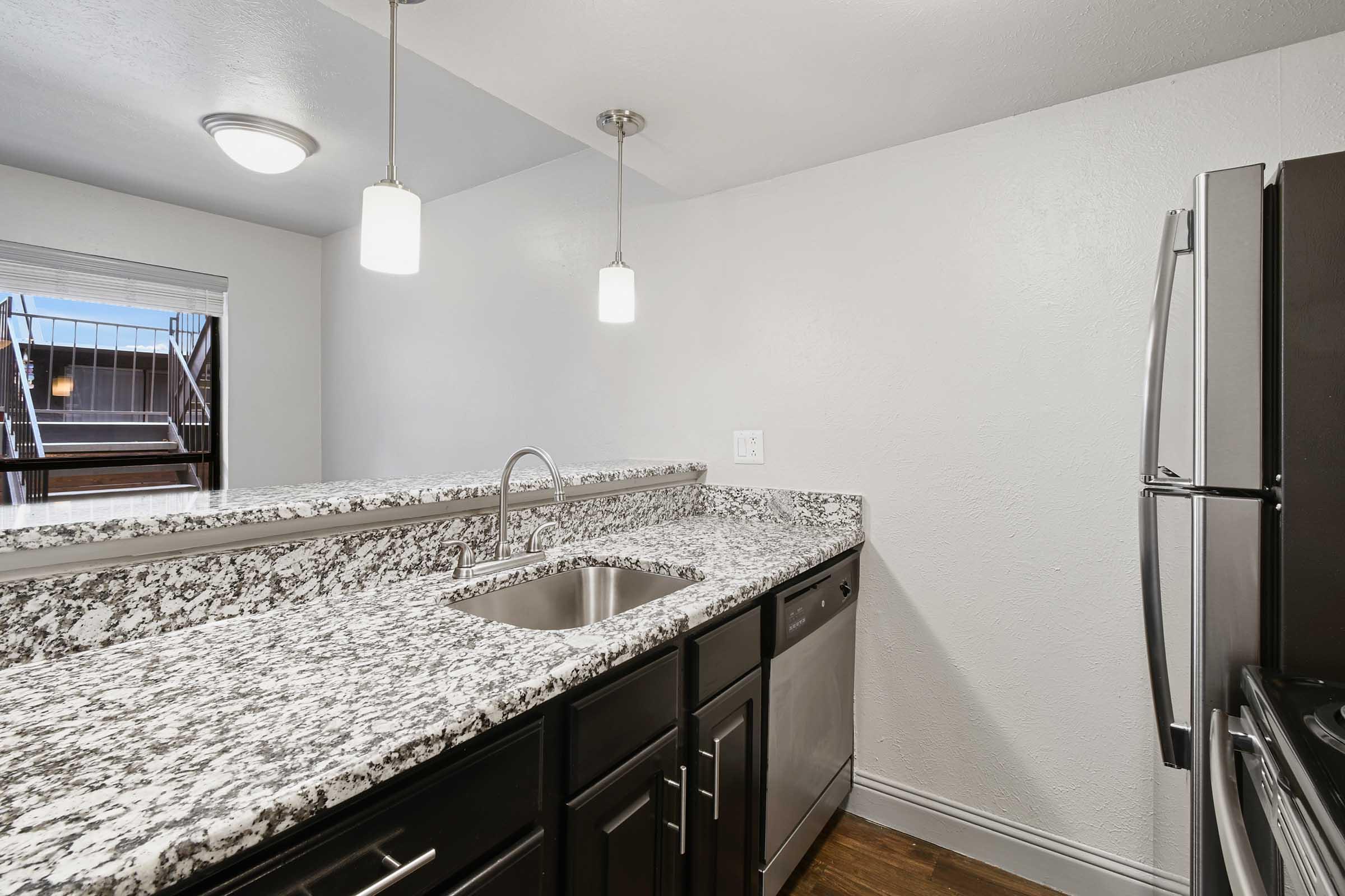 Modern kitchen featuring a granite countertop, stainless steel sink, and dark cabinets. The space includes pendant lighting above the counter and a window providing natural light. Stainless steel appliances, including a dishwasher, are present. The warm wood floor adds to the inviting atmosphere.