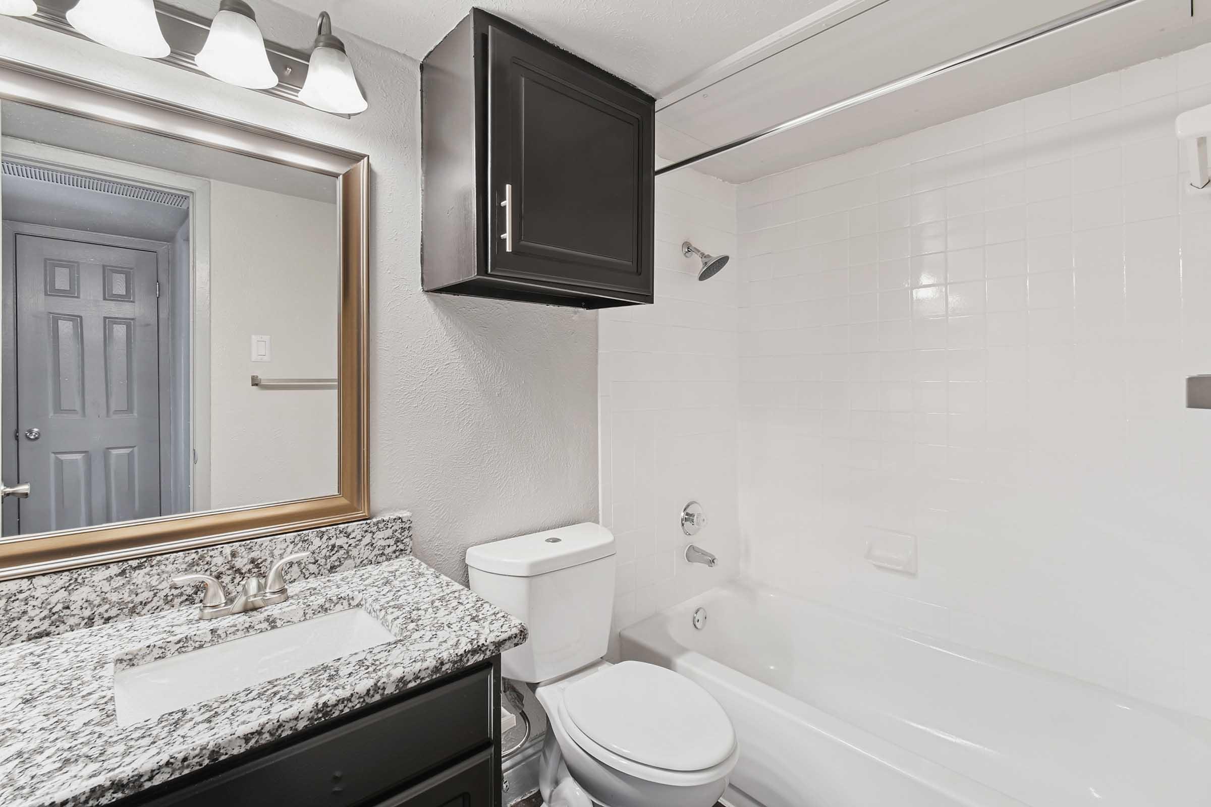 A modern bathroom featuring a white bathtub, a toilet, and a granite countertop with a sink. There is a large mirror above the sink, and stylish lighting fixtures. The walls are tiled, and there is a door visible in the background.