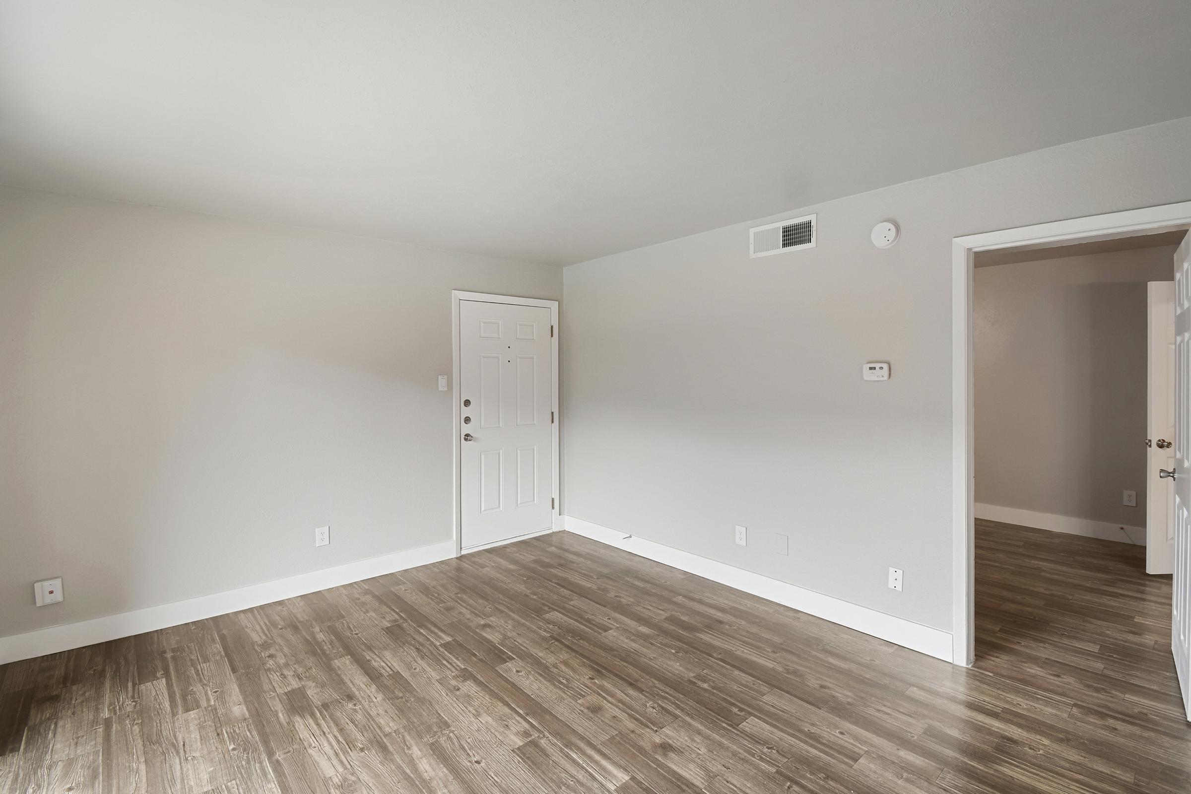 A spacious, empty room with light gray walls and wooden flooring. A white door is visible on the left side, and a closet door is seen to the right. Natural light fills the space, making it bright and airy. The room is unfurnished, highlighting its potential for decoration and personalization.