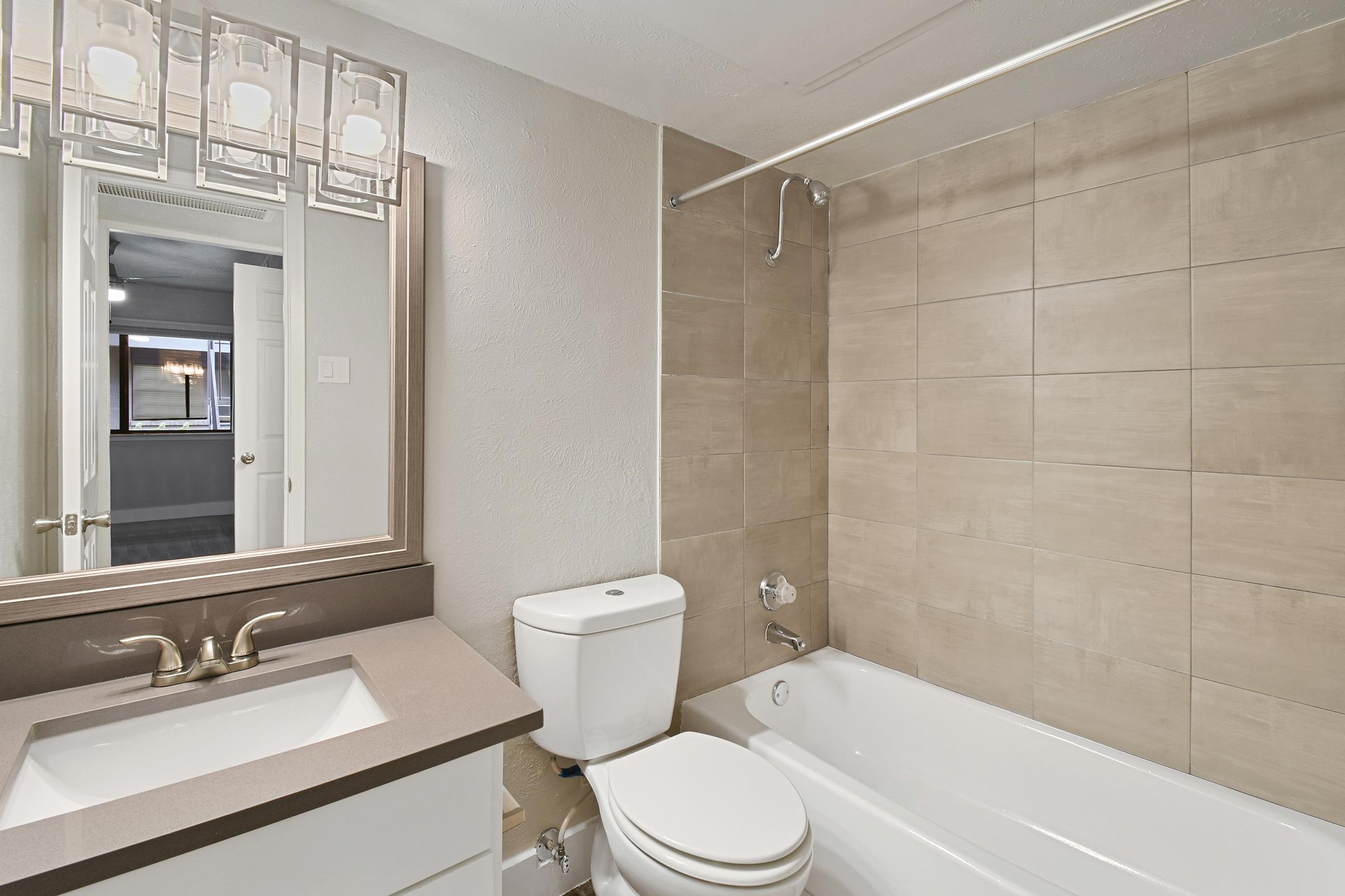Modern bathroom featuring beige tiled walls, a bathtub with a shower, a white toilet, and a sleek vanity with a mirror. Four light fixtures hang above the mirror, providing soft illumination. The space is clean and contemporary, with a door leading to an adjacent room visible in the mirror's reflection.