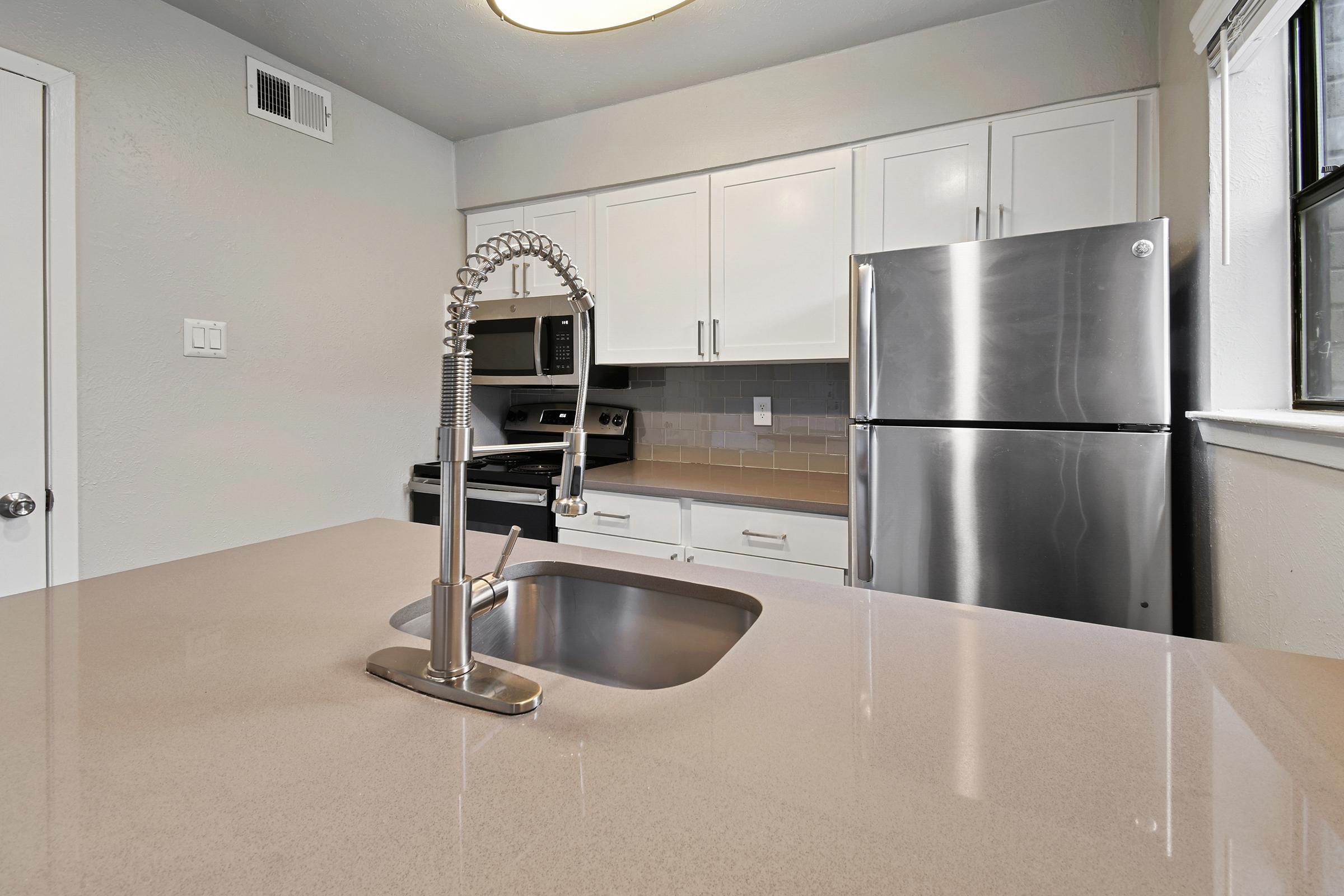 Modern kitchen featuring a stainless steel refrigerator, stove, and microwave. The countertop is smooth and light-colored, with a sleek sink and a stylish faucet. Cabinets are white, creating a bright and clean space, with a window providing natural light.