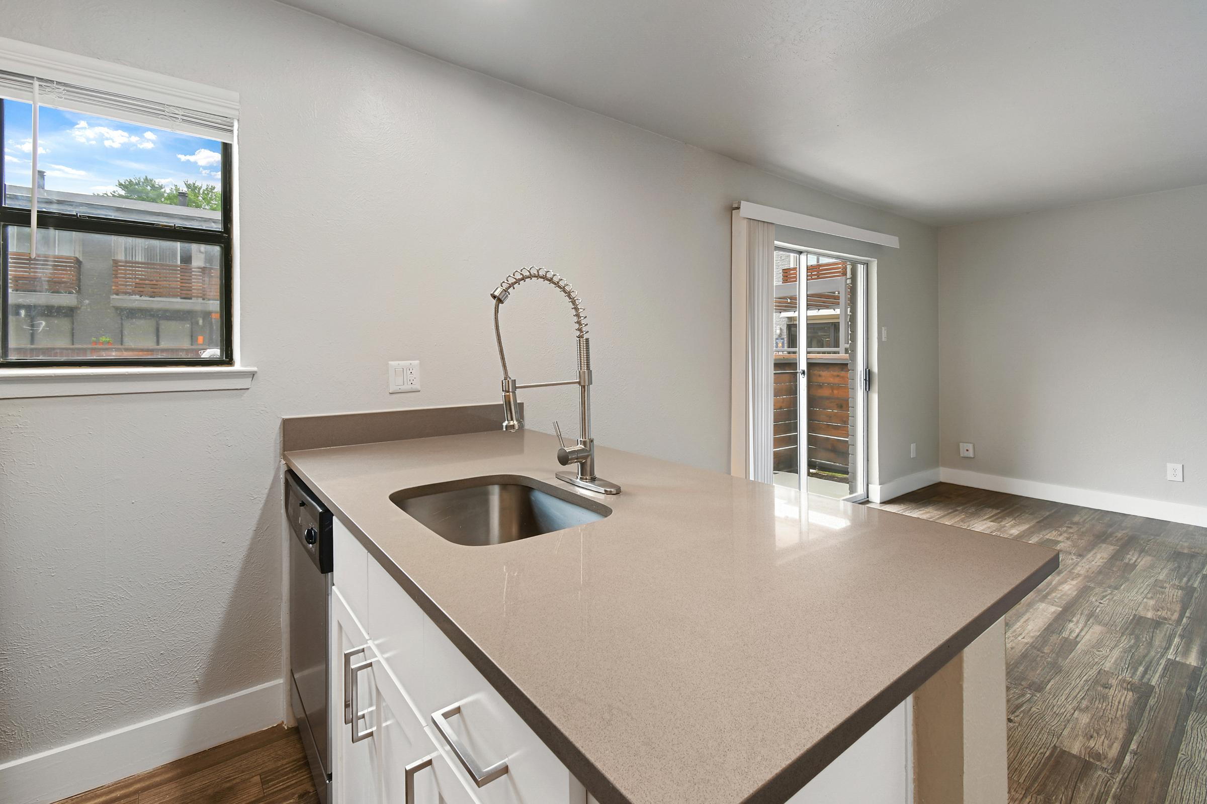 A modern kitchen with a clean countertop, sink, and a chrome faucet. The space features light-colored walls and large windows allowing natural light, with a view of a patio or balcony. The flooring is wooden, and there is an open area leading to a living space.