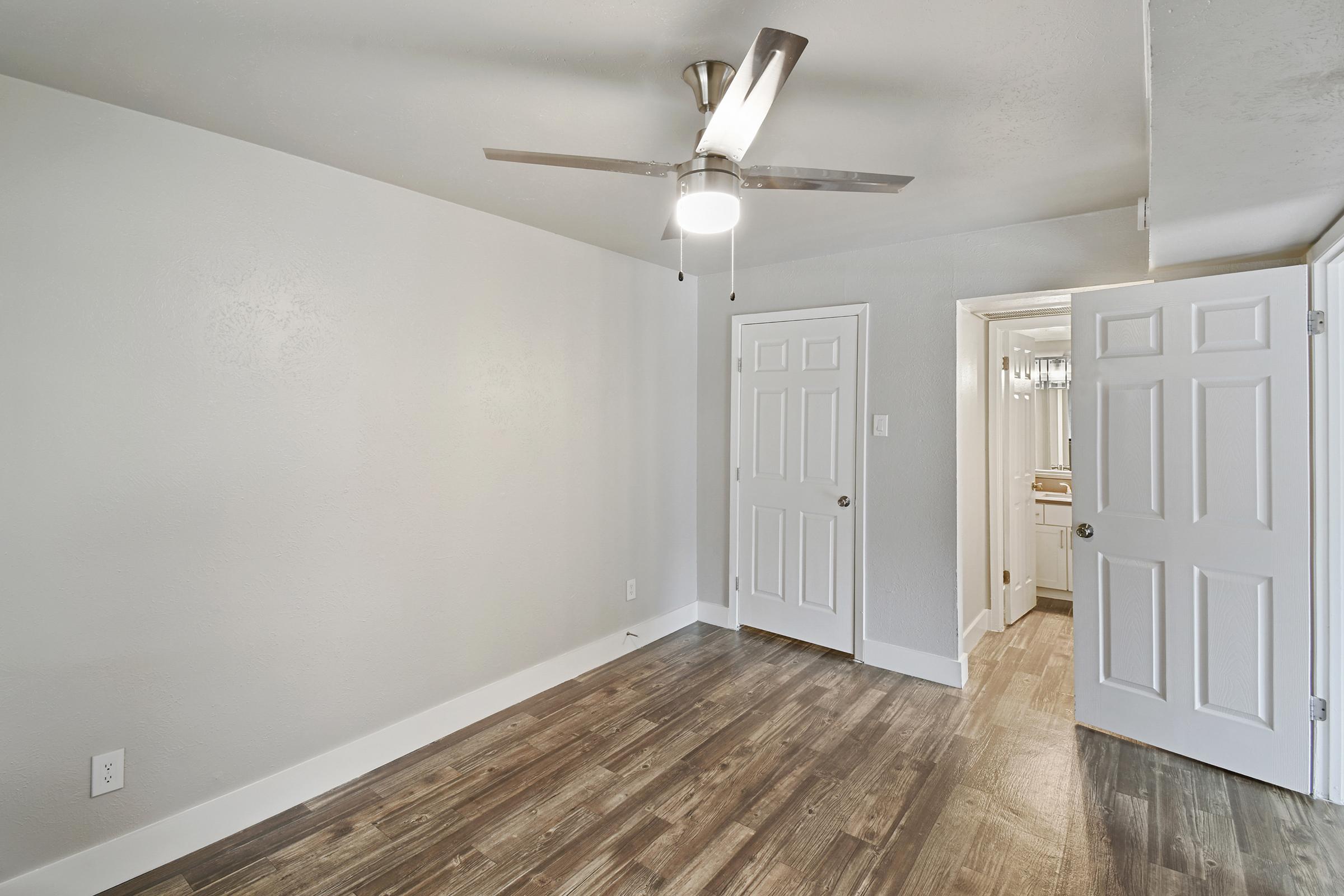 A spacious room featuring a ceiling fan, light-colored walls, and hardwood-style flooring. Two white doors lead to a closet and a bathroom. The room is empty, providing a bright and fresh atmosphere with ample natural light.
