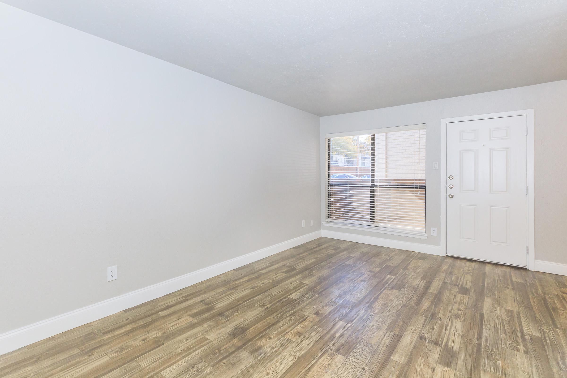A spacious, empty living room with light-colored walls and a large window allowing natural light. The floor is covered in wooden planks, and there is a white door leading outside. The room has a clean, minimalist look, perfect for various design options.