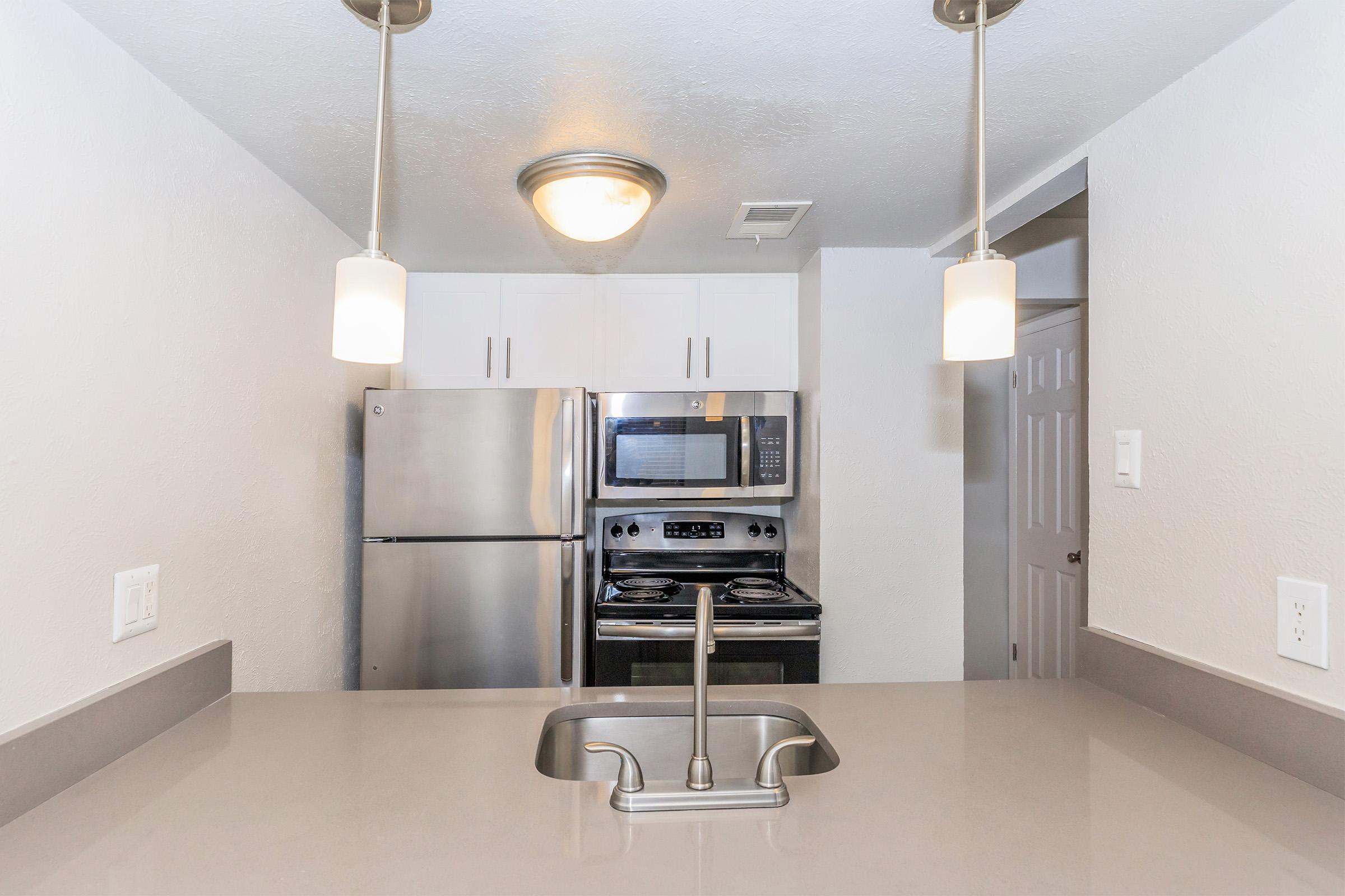 Modern kitchen with stainless steel appliances, including a refrigerator, microwave, and stove. The countertop is sleek and gray, with a sink in the center. Two pendant lights hang above, illuminating the space. White cabinets line the walls, creating a bright and clean atmosphere.