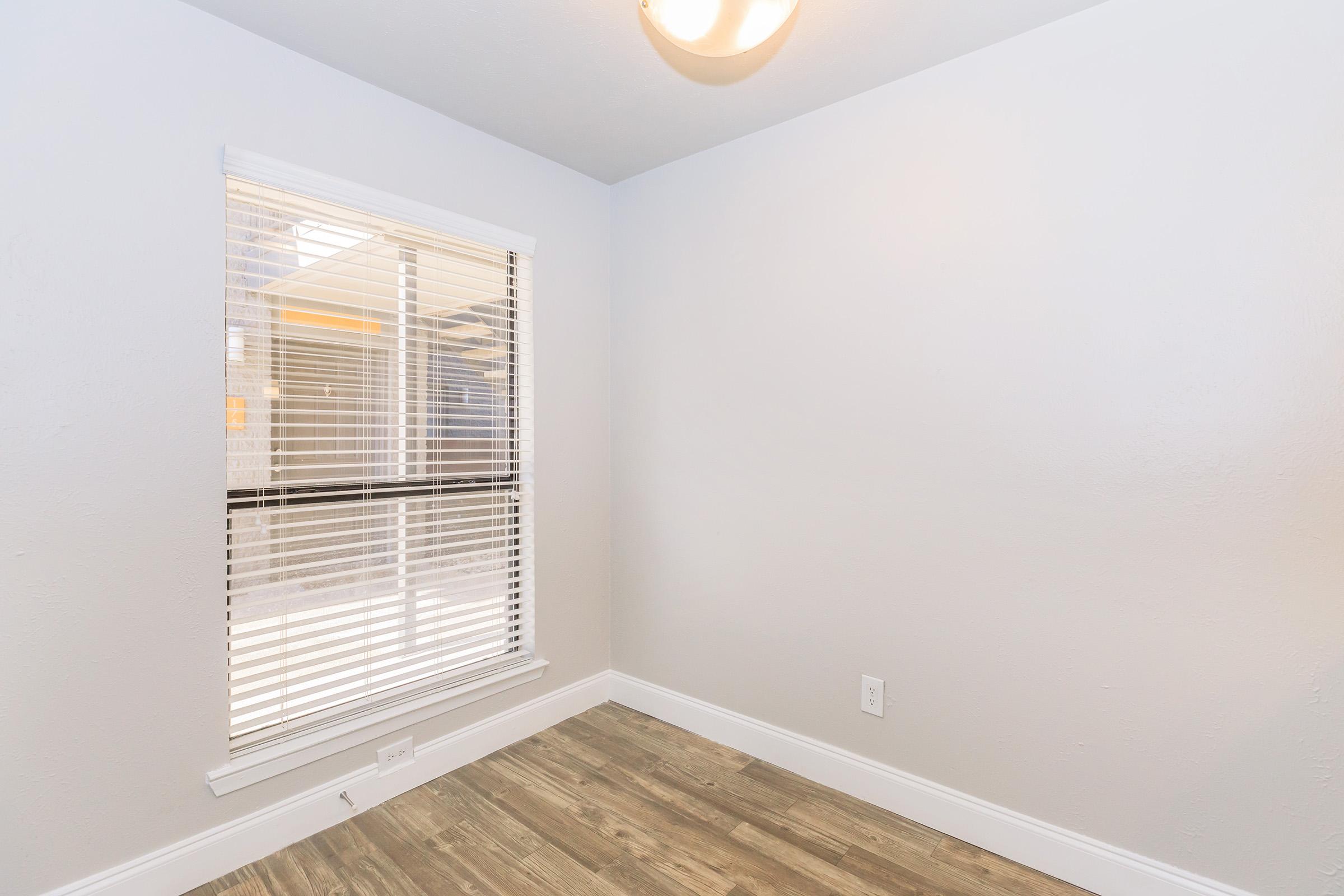 A small, empty room with light gray walls and wooden flooring. There is a window with white blinds allowing natural light, and a ceiling light fixture. The room has a minimalist design, making it suitable for various uses.