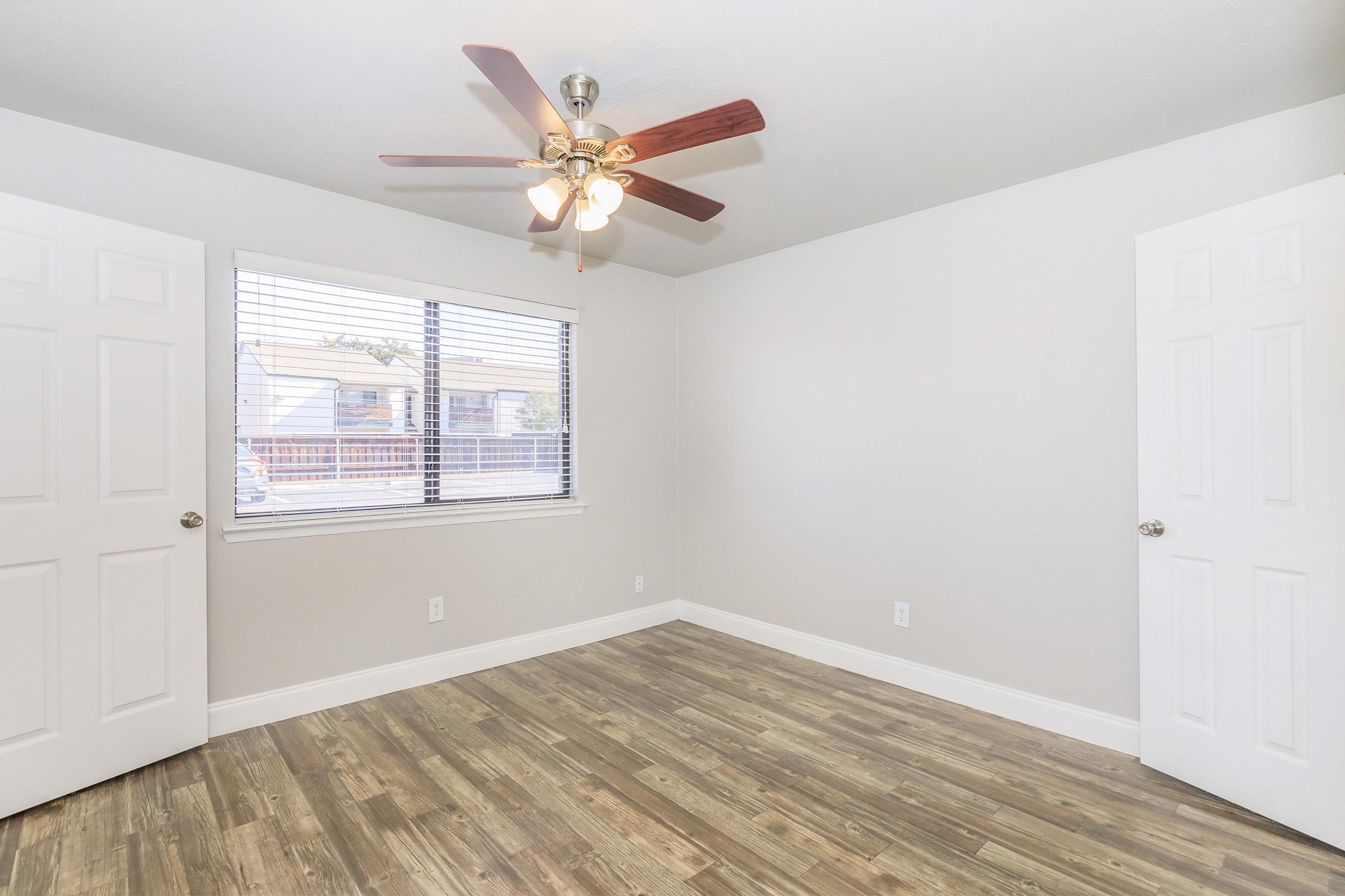 A bright, empty room featuring a ceiling fan with light fixtures, two white doors, and a large window with blinds. The floor is adorned with wood-like laminate, and the walls are painted in a soft, neutral color, creating a clean and inviting ambiance.