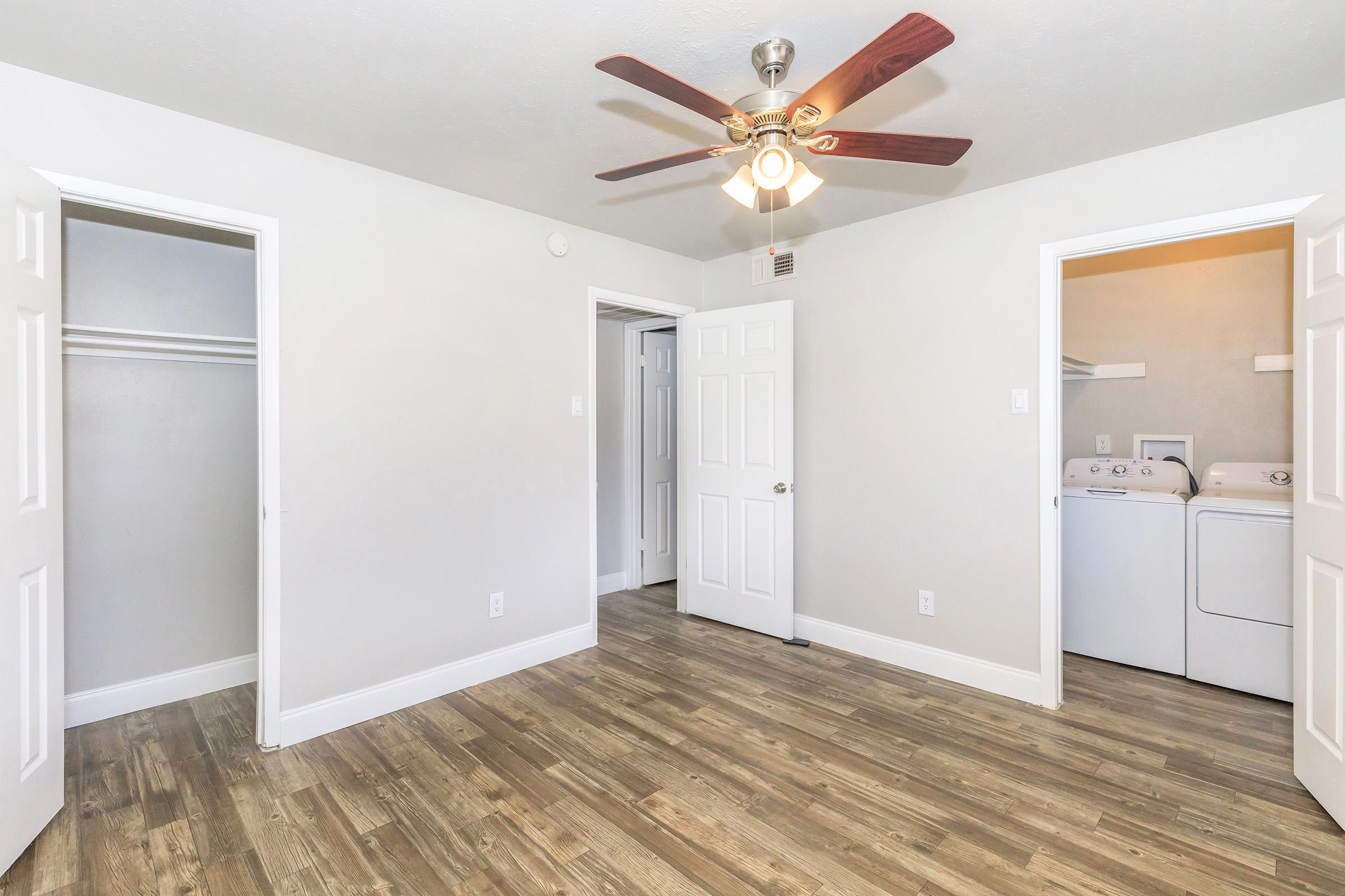Empty room with light-colored walls and a ceiling fan. Two doors lead to adjacent spaces, one possibly a closet and the other a laundry area with stacked washer and dryer. The floor has a wood-like finish, creating a warm atmosphere.