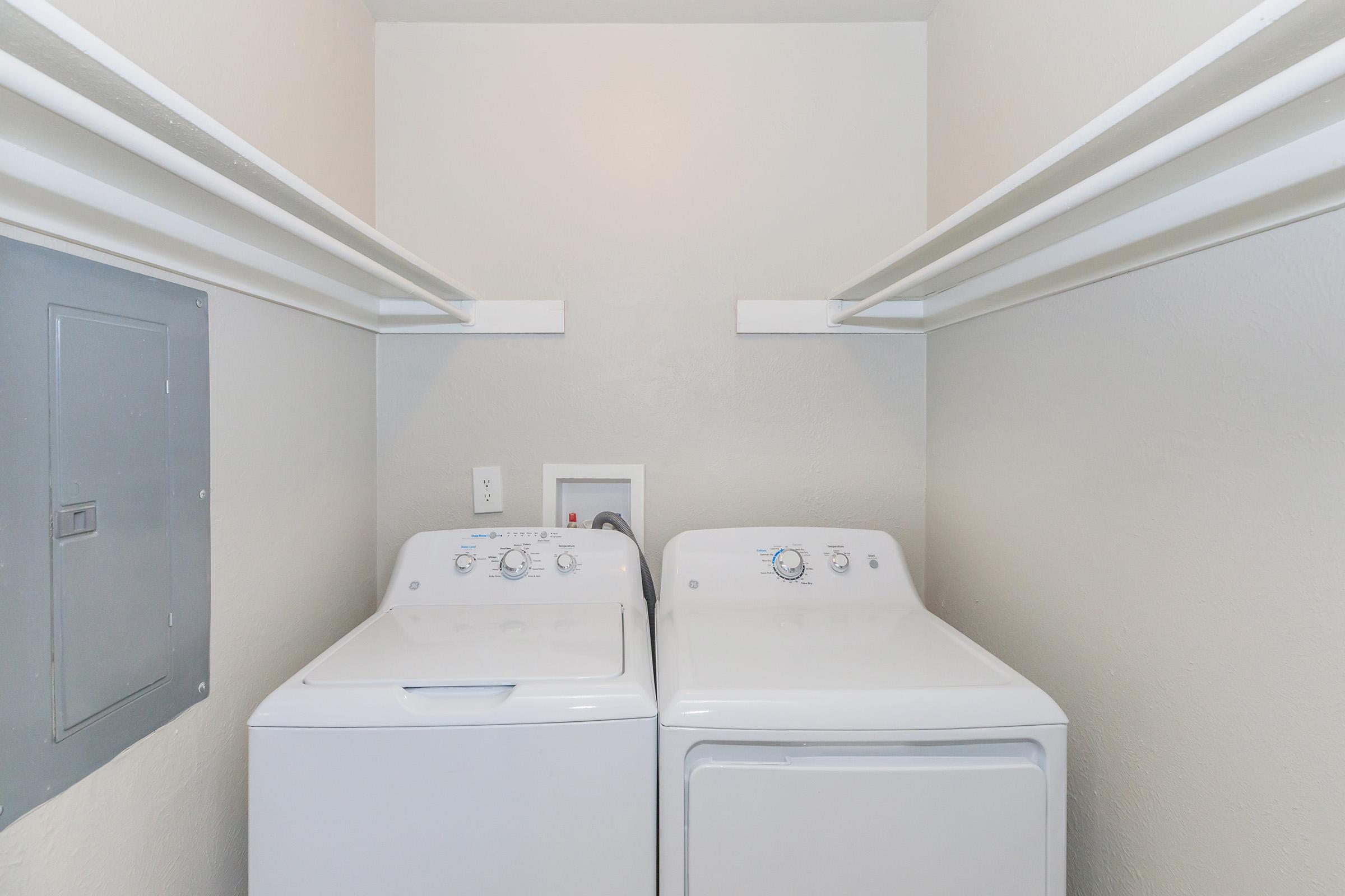 A laundry room featuring a white washing machine and dryer side by side. The walls are light-colored, and there are two shelves above the appliances for storage. An electrical panel is visible to the left. The overall space appears clean and organized.
