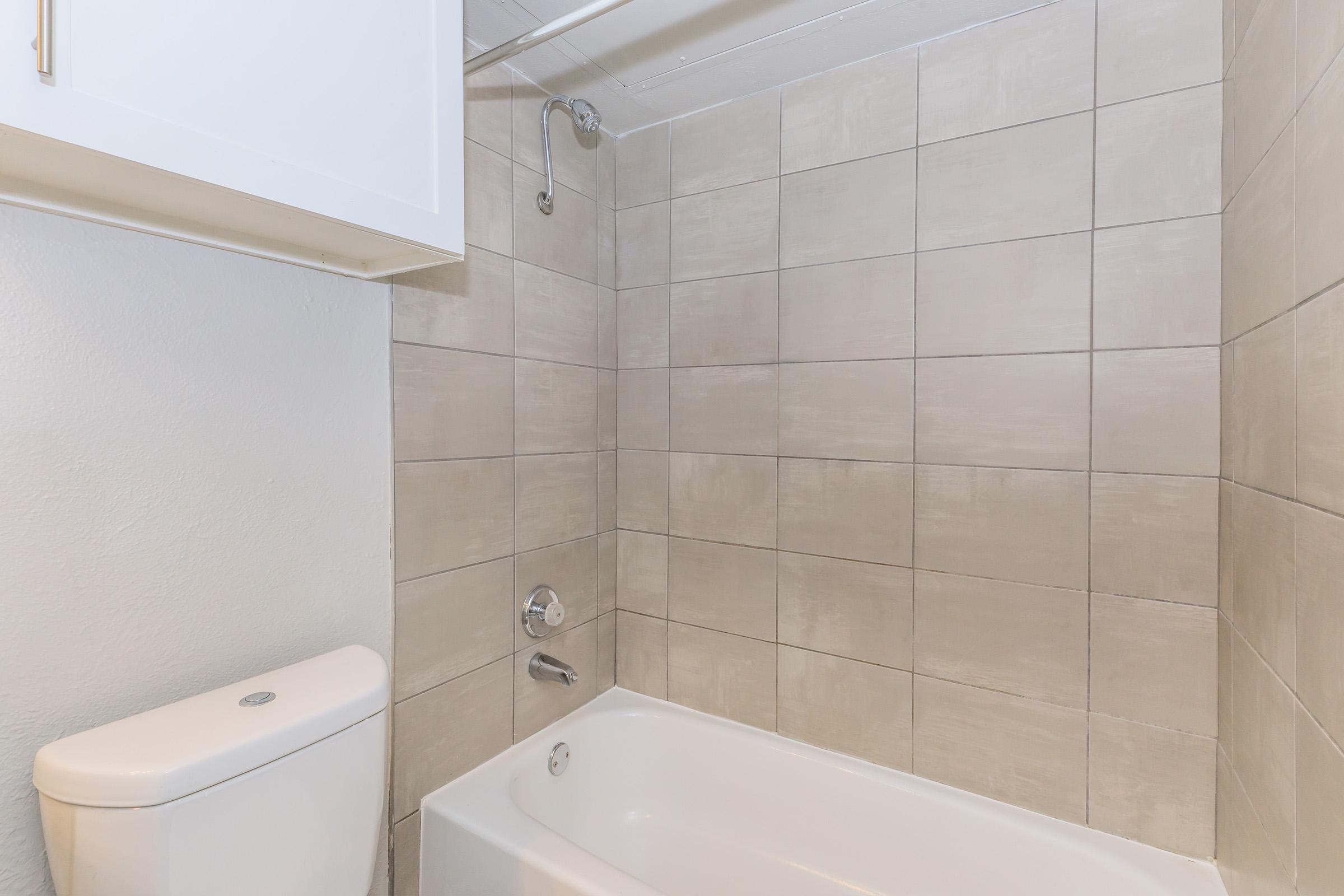 A clean bathroom featuring a bathtub with a showerhead above, tiled walls in neutral shades, and a white toilet. The space is well-lit and minimalistic, with a cabinet above the bathtub for storage.