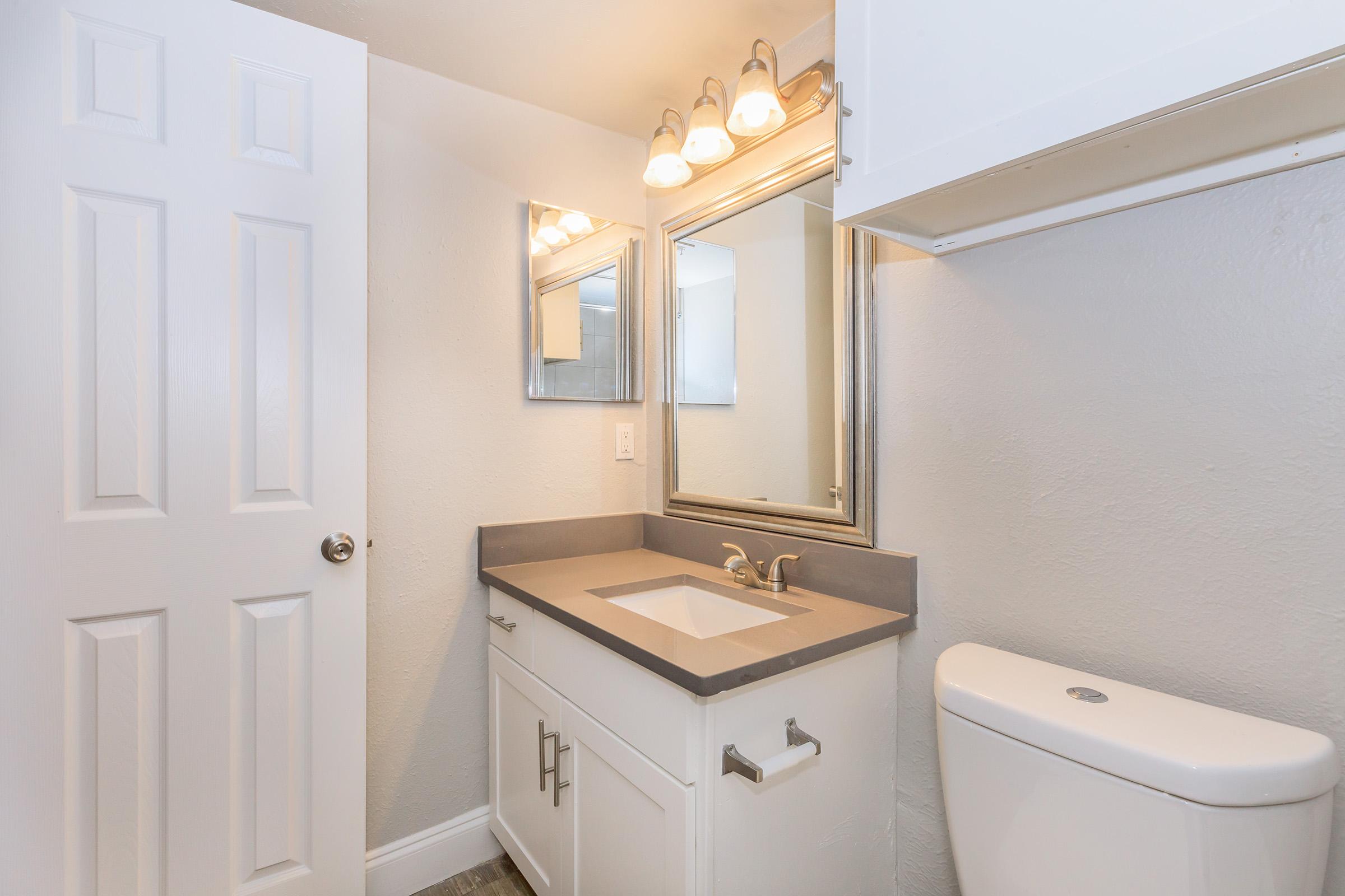 A tidy bathroom featuring a white vanity with a gray countertop, a rectangular sink, and modern chrome fixtures. Above the sink, there are two mirrors and four light fixtures. The wall is painted light gray, and there is a white toilet beside the vanity. A closed white door is visible on the left.