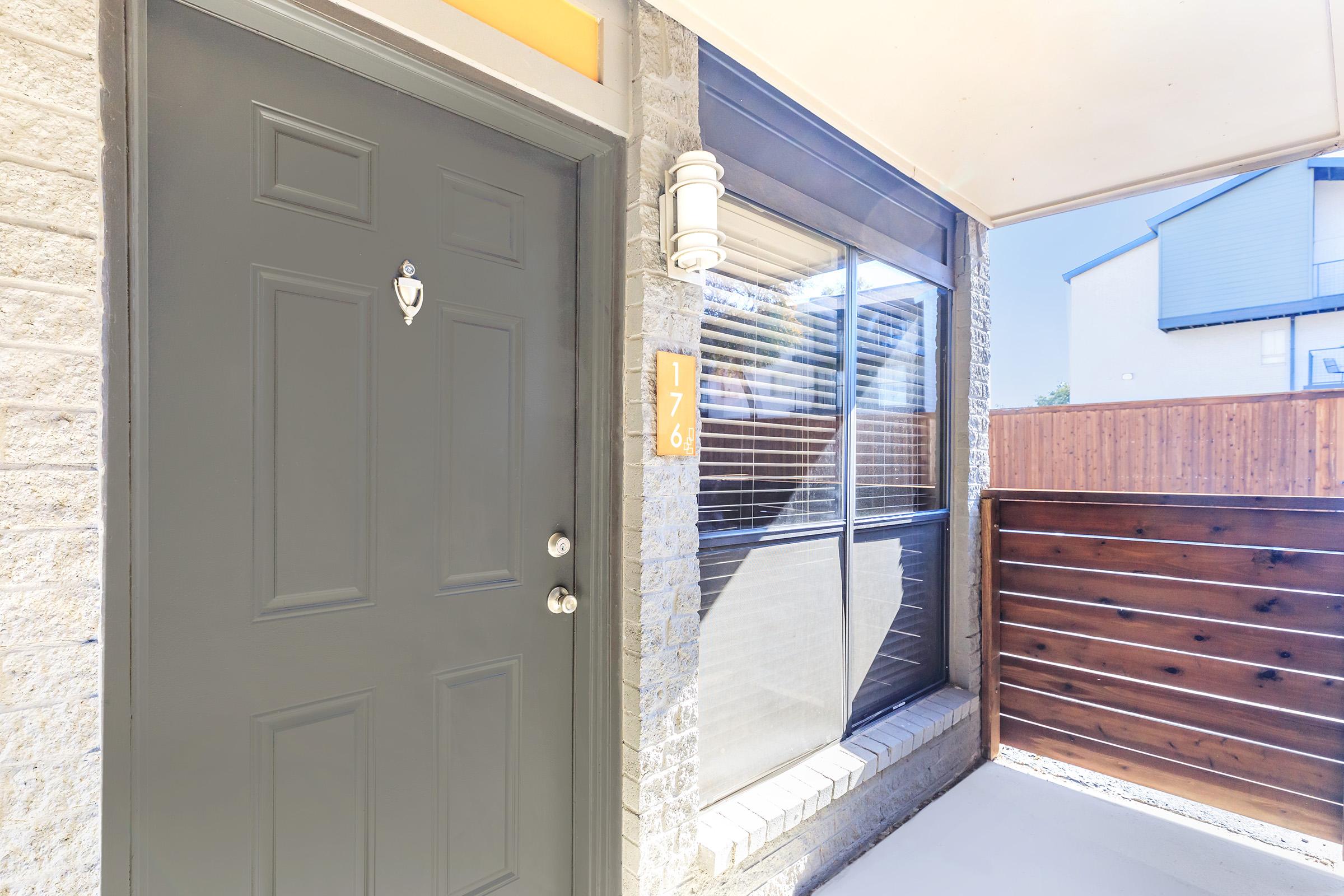 Close-up of a modern entrance door with a dark finish, featuring a doorbell and a house number '176' displayed on the side. The door is framed by light-colored stone and a wooden fence is visible in the background, with windows showing a view of the outdoors. Bright natural light illuminates the scene.